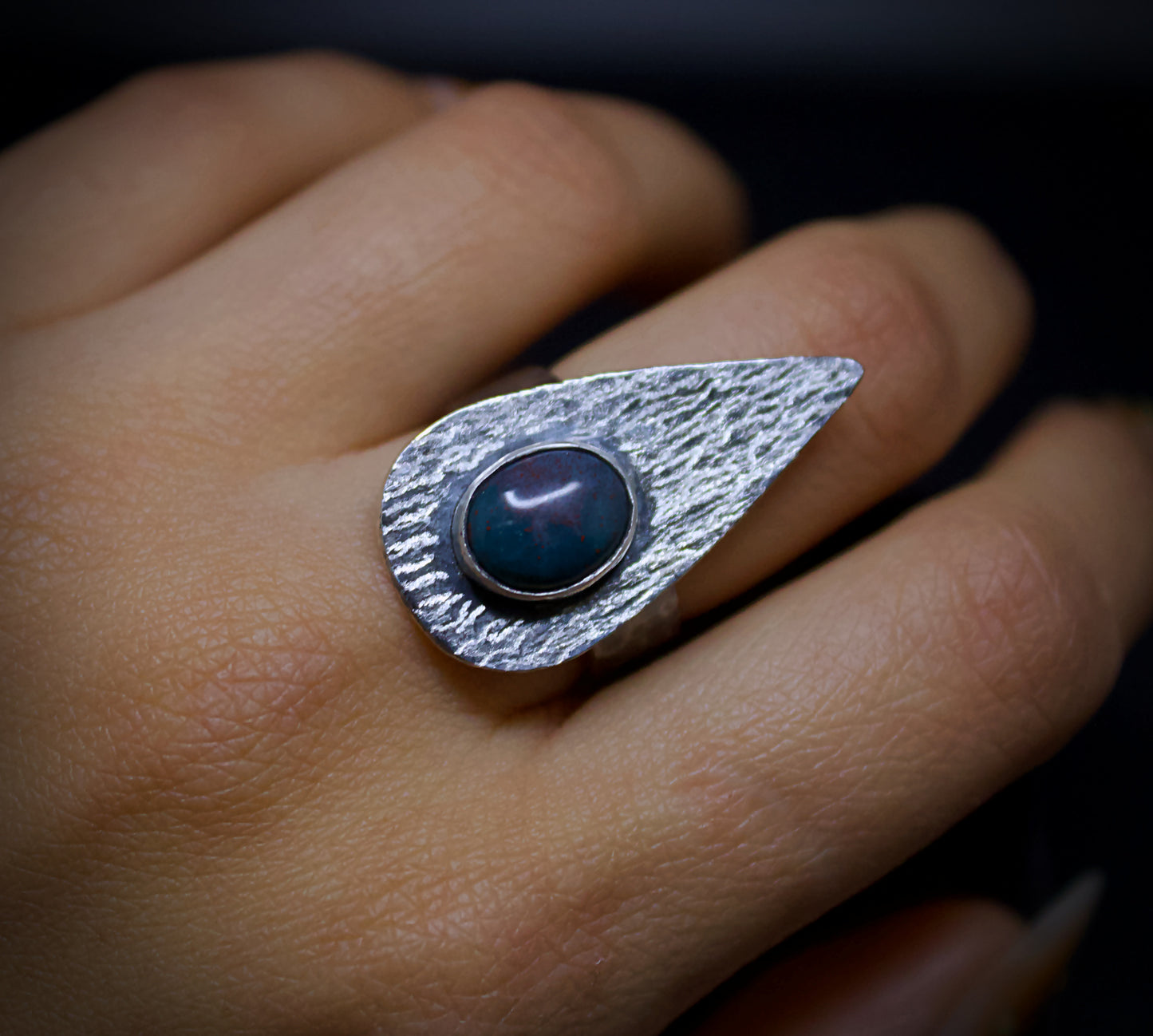 Silver ring with a green and red gemstone on a finger against a dark background