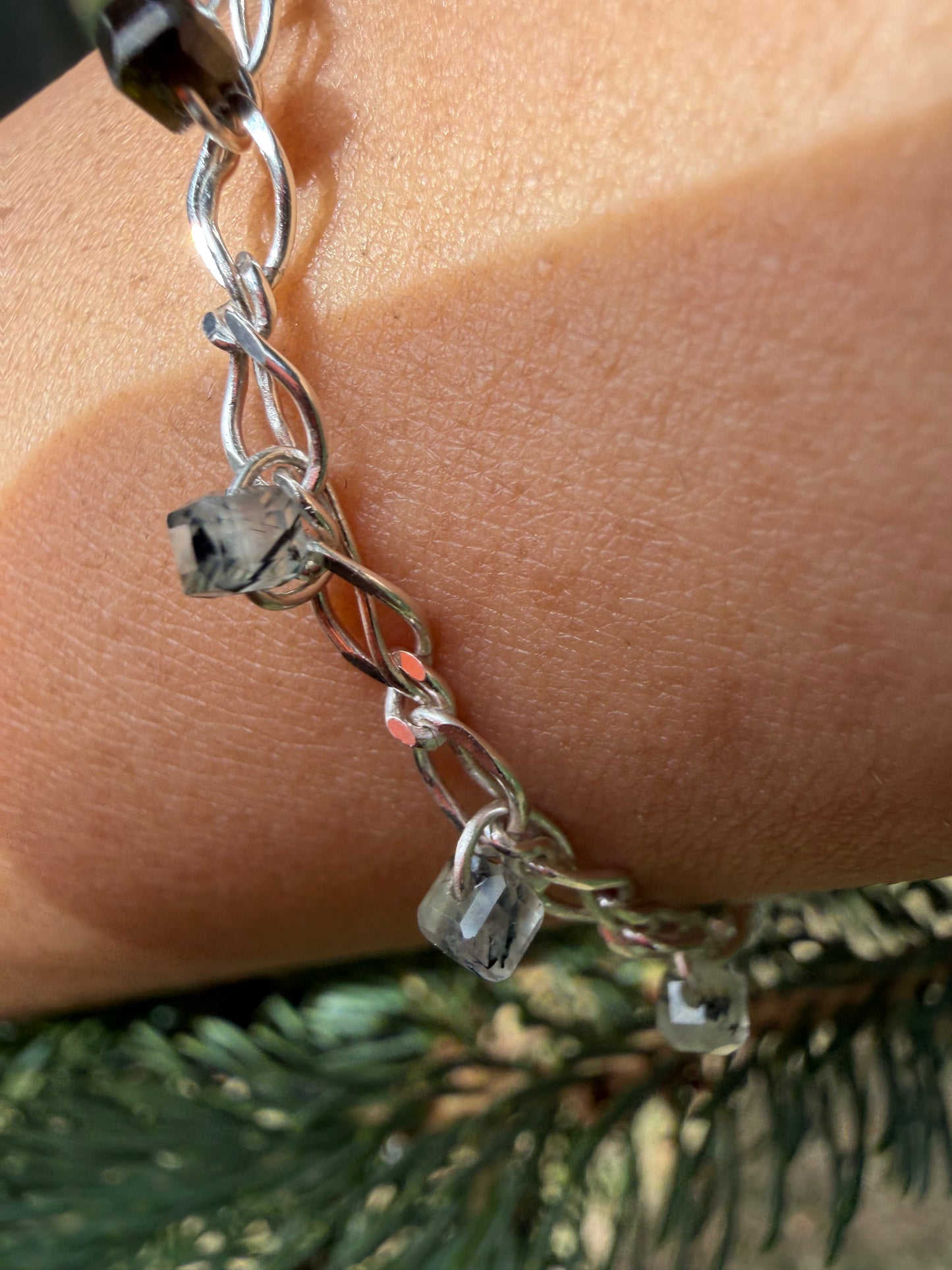 A close-up photograph of the handcrafted Lumos Bracelet worn on a wrist, positioned against a background of natural green pine needles. The bright, shiny sterling silver chain catches the light, showcasing translucent, ice-like prehnite stone beads threaded onto the links.