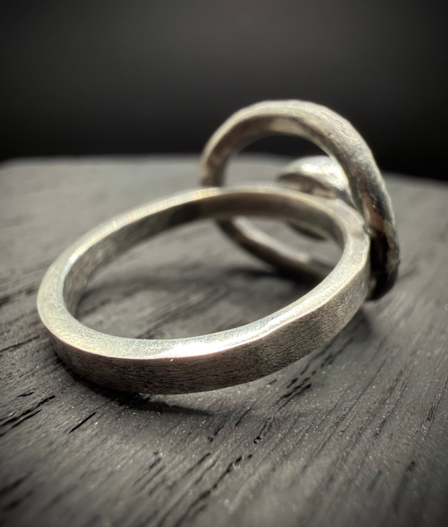 A side profile view of a handcrafted sterling silver ring resting on a dark wood surface, showcasing a smooth, uniform band that connects to a prominent, textured open circle frame.