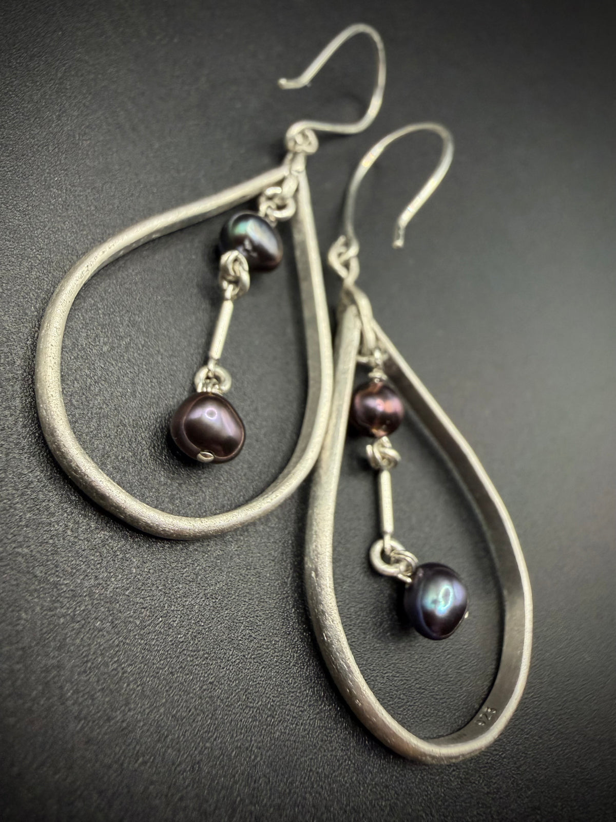 A pair of silver teardrop dangle earrings with small iridescent dark purple beads, displayed on a dark textured background.