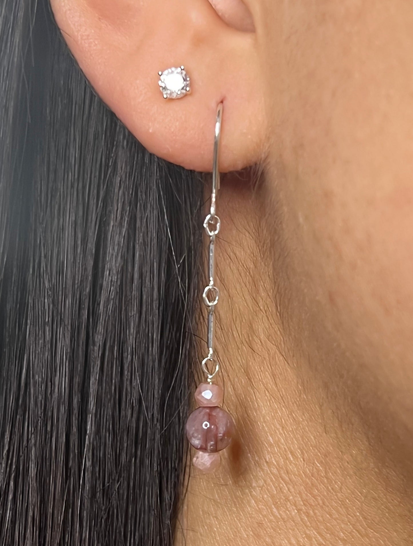 A close-up of a person wearing a handcrafted sterling silver earring featuring a long delicate chain with a stack of faceted peach moonstone and smooth red agate beads.
