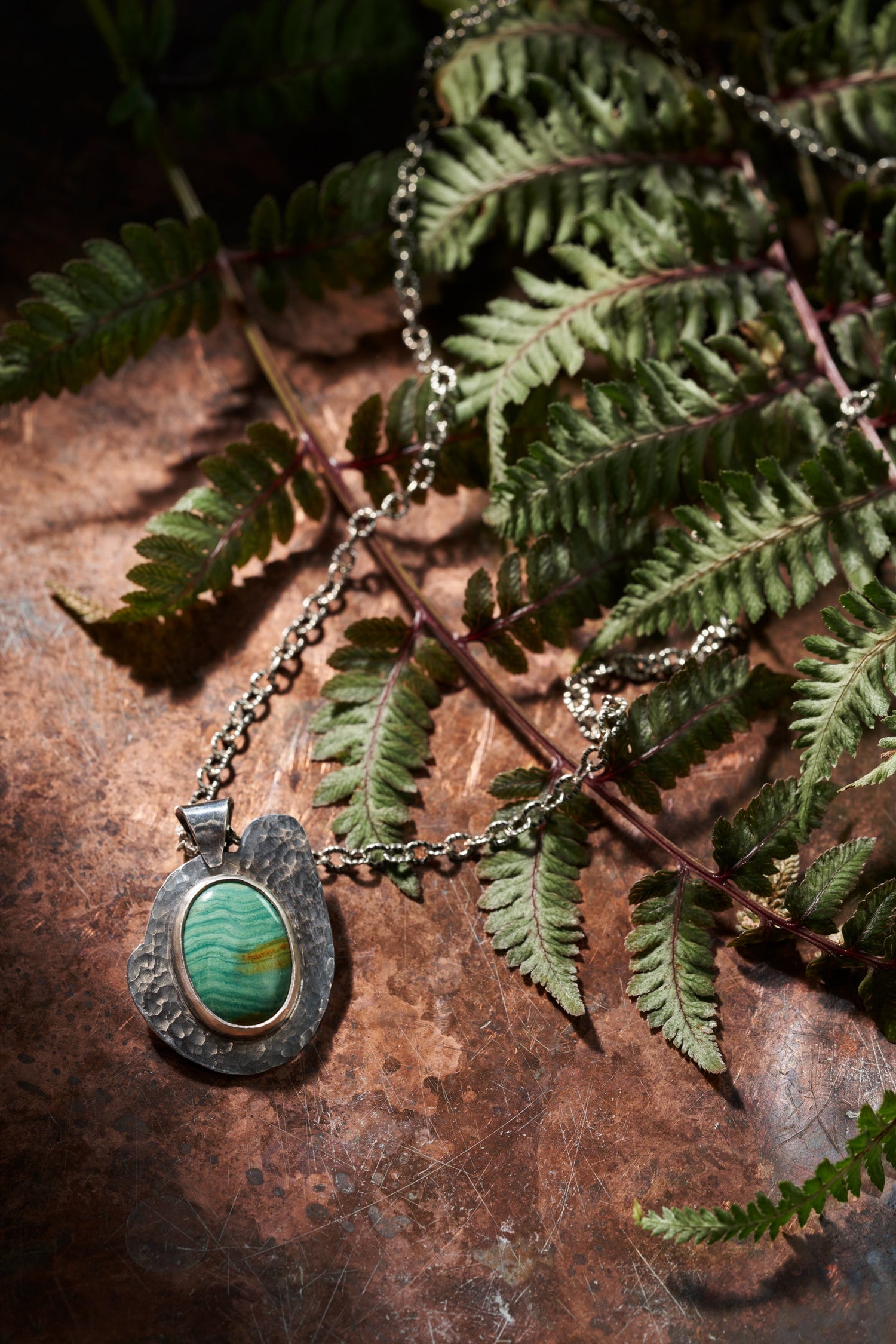 Textured silver pendant with a green stone. Displayed on a natural brown surface and surrounded by fern like leaves. 