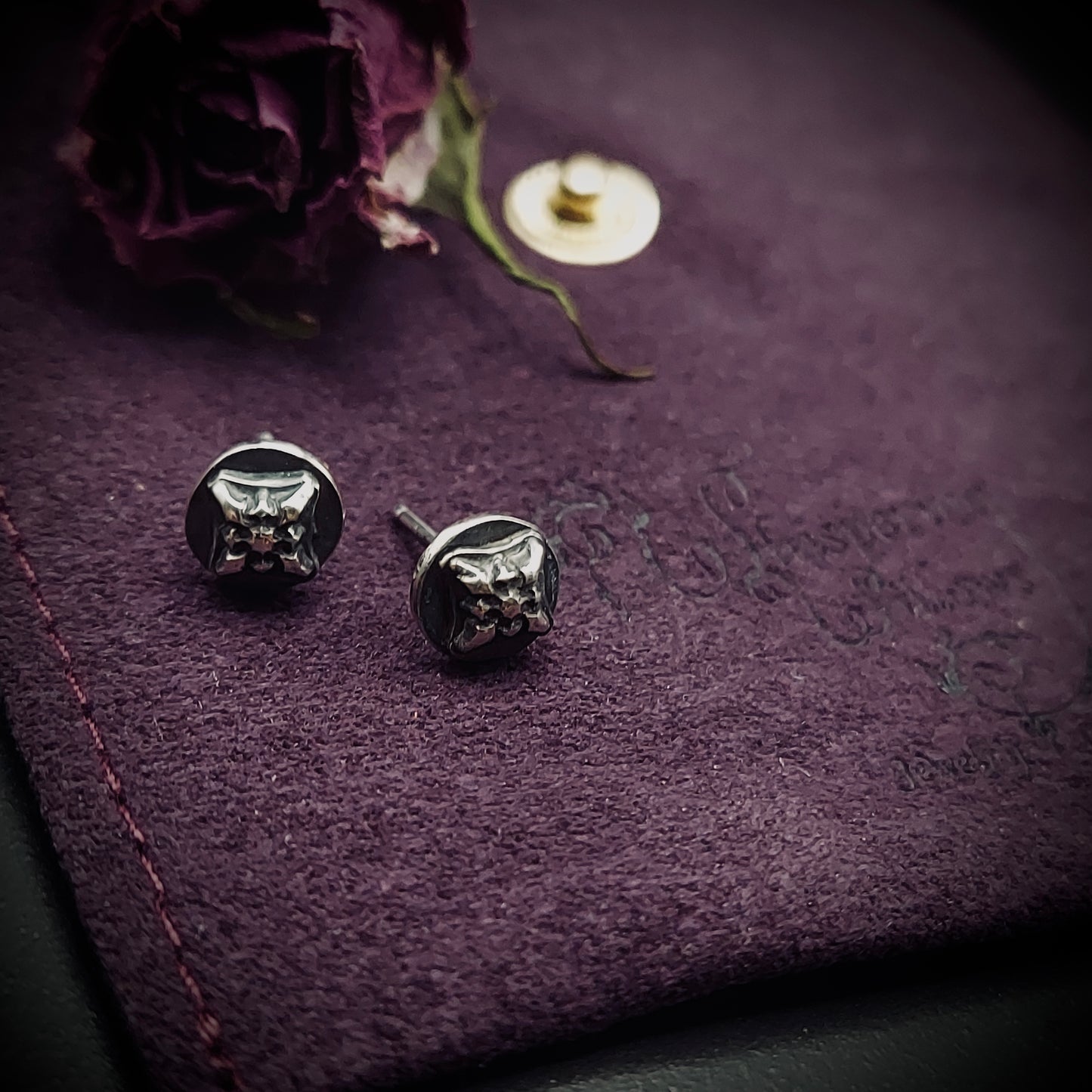 Sterling silver Ember Stud earrings from the Ashborn Collection. The studs feature a raised, stylized cat head design with a polished finish, contrasted against a dark, recessed patina. They are displayed on a dark purple textured background next to a dried rose.