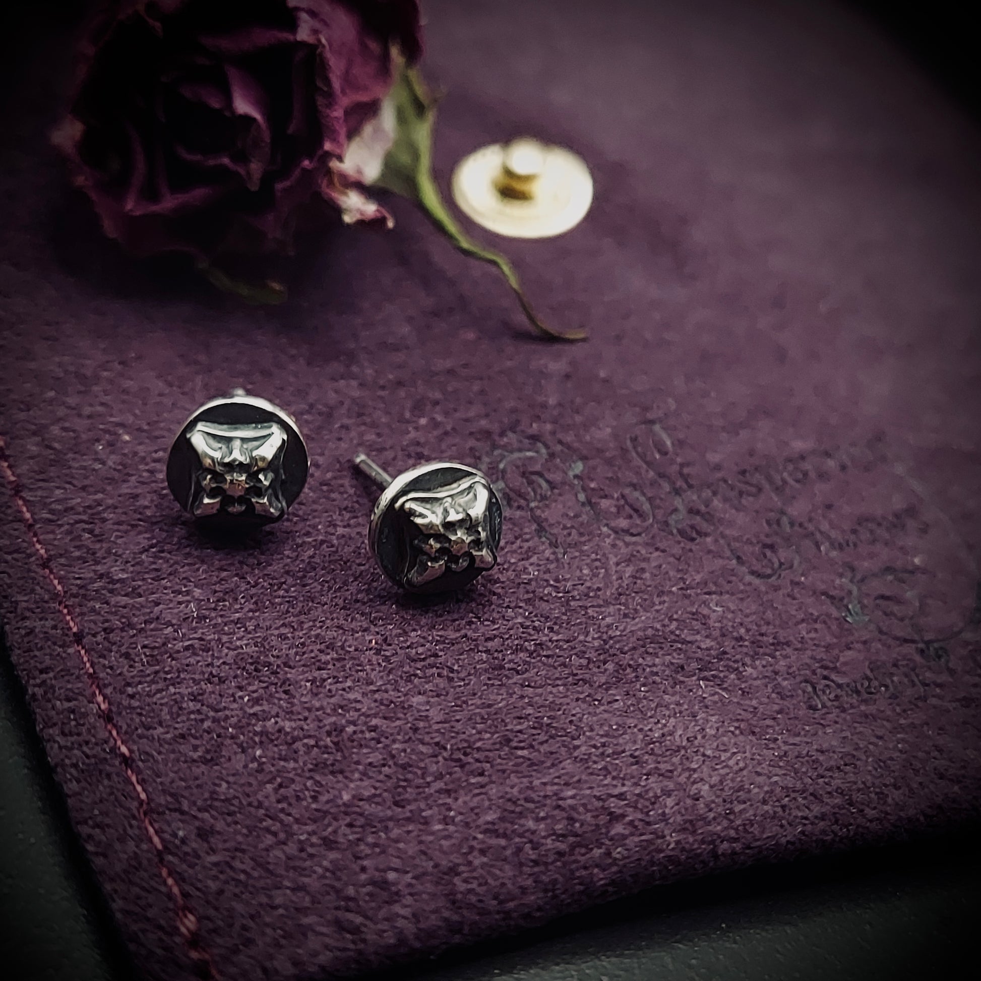 Sterling silver Ember Stud earrings from the Ashborn Collection. The studs feature a raised, stylized cat head design with a polished finish, contrasted against a dark, recessed patina. They are displayed on a dark purple textured background next to a dried rose.