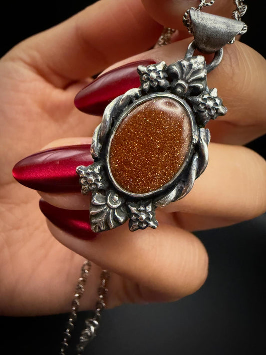 Silver pendant with brown stone held by a hand with red nail polish against a dark background