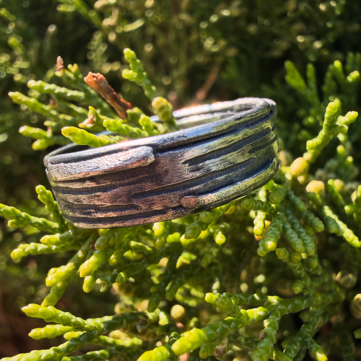 Handcrafted sterling silver Rune Ring with a textured wood-grain finish resting on green conifer branches.