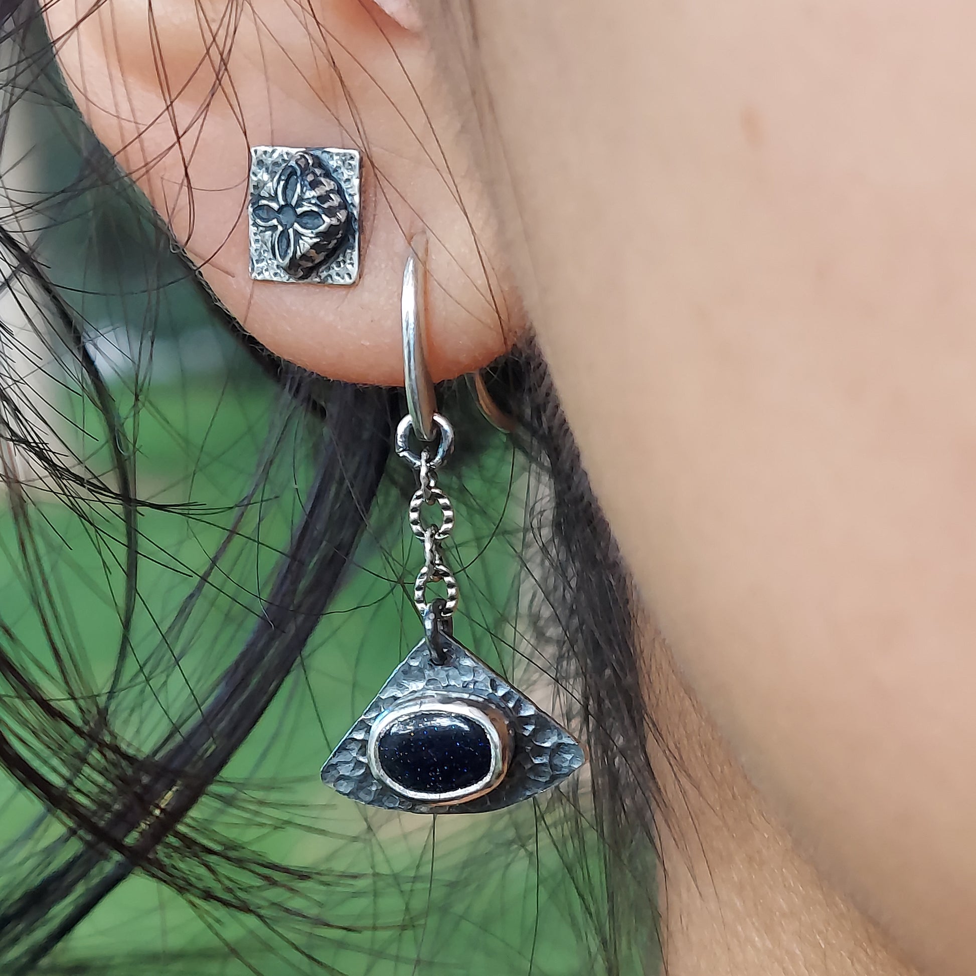 Close-up of Astraea Drop earrings on a dark, moody background, highlighting the sparkling galaxy-like blue goldstone and the weathered, hammered sterling silver texture.