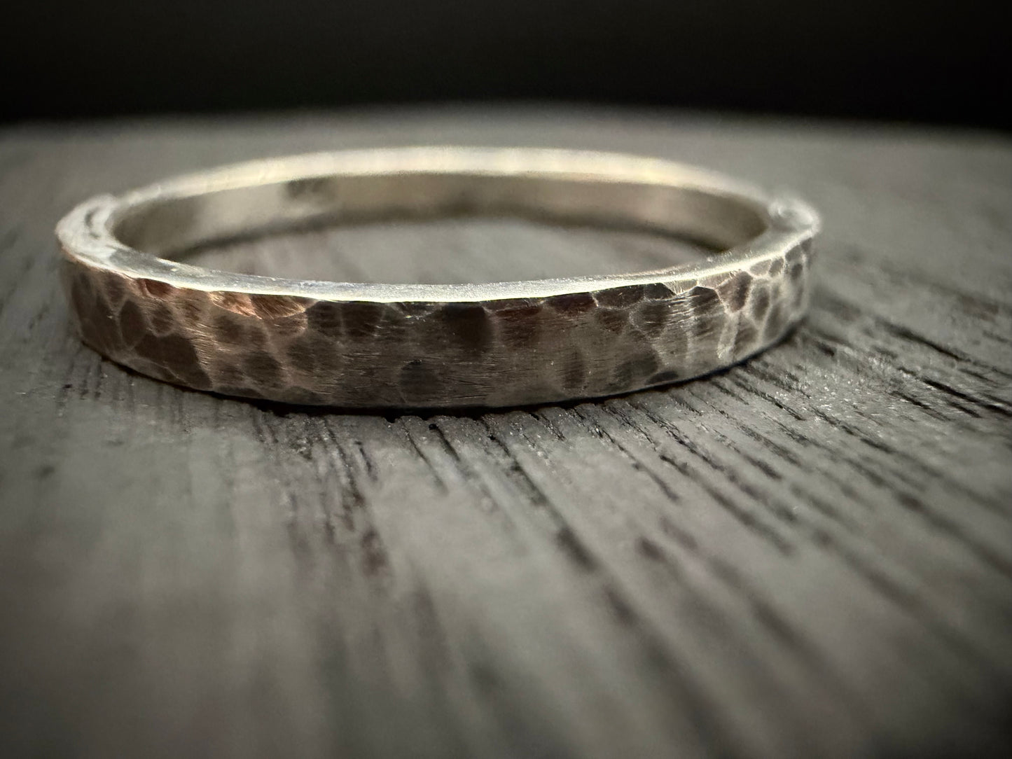 A close-up photograph of a handcrafted sterling silver band featuring a hammered texture. The ring, identified as the Blackwood Band, sits on a textured wooden surface. 