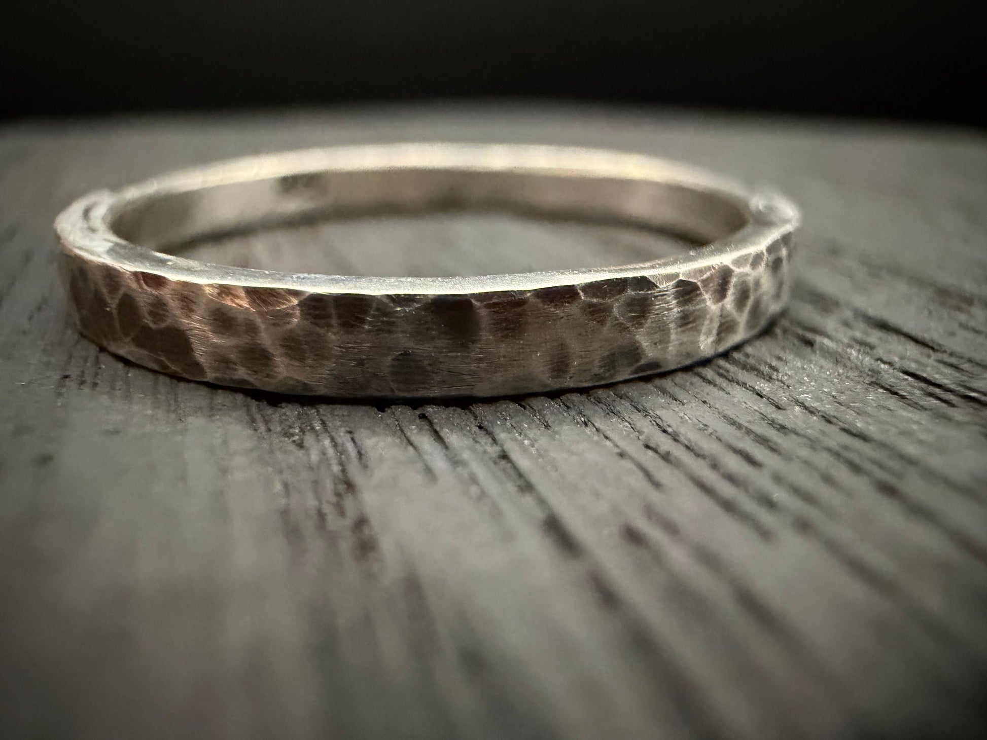 A close-up photograph of a handcrafted sterling silver band featuring a hammered texture. The ring, identified as the Blackwood Band, sits on a textured wooden surface. 