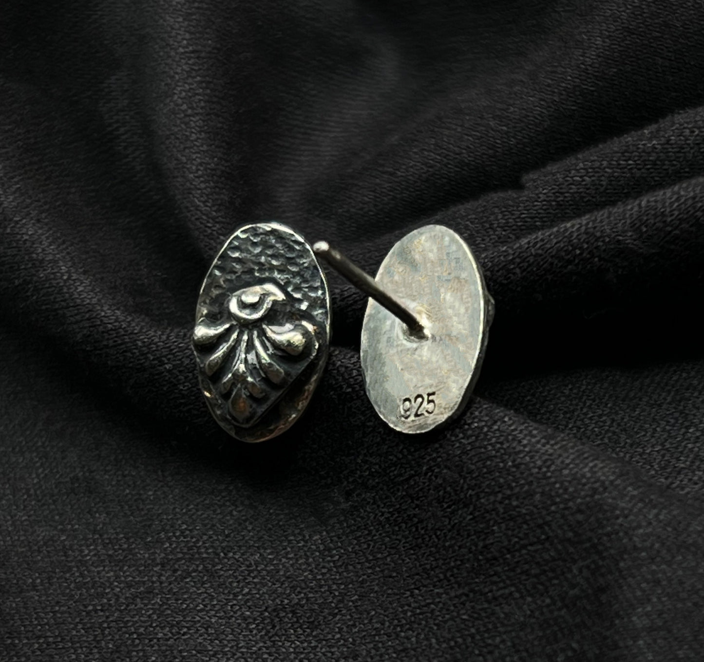 Sterling silver oval earrings resting on dark textured fabric. The high-contrast oxidation mimics soot and shadow, emphasizing the structural, tiered design and the hand-wrought details of the metal.