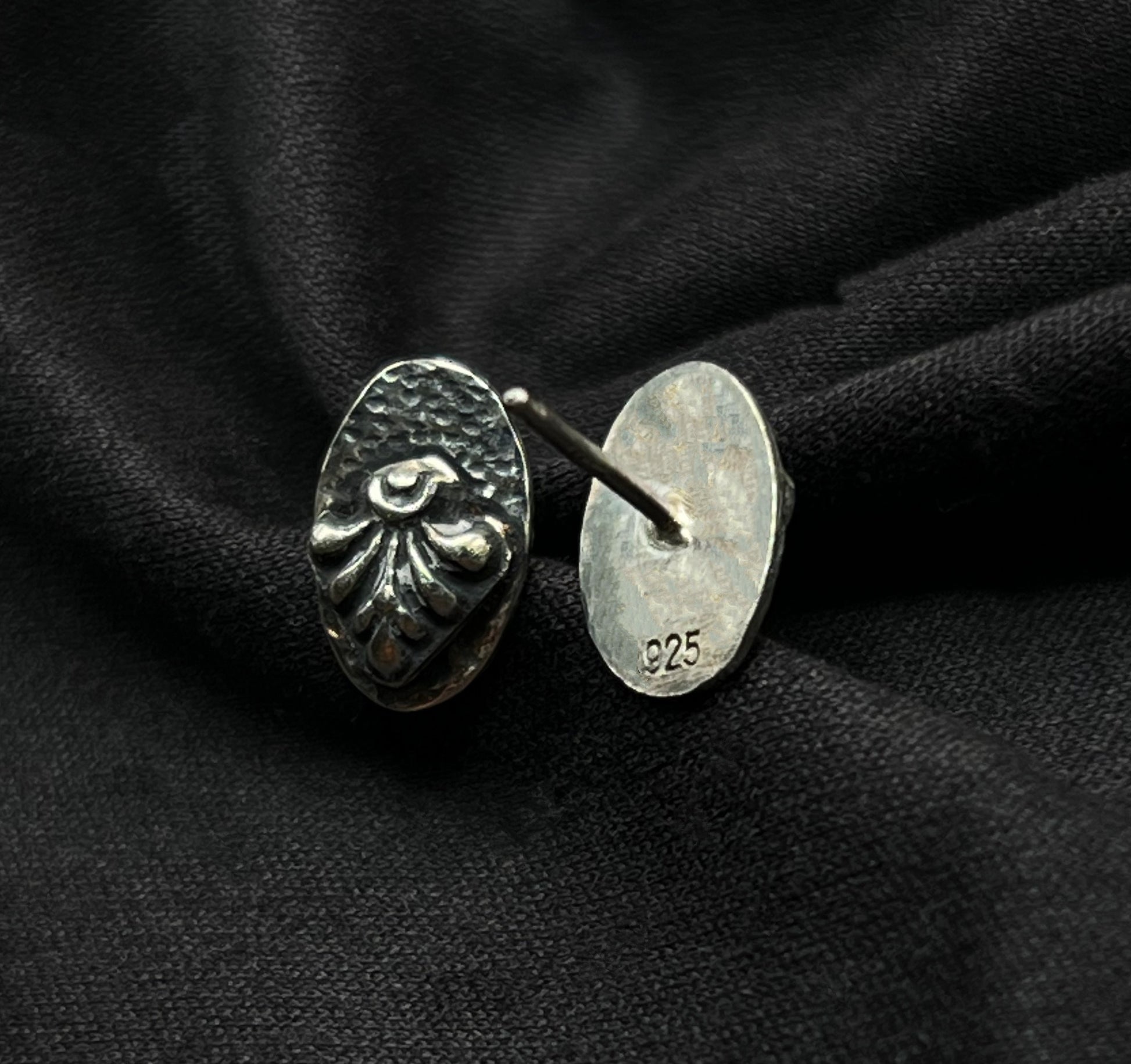 Sterling silver oval earrings resting on dark textured fabric. The high-contrast oxidation mimics soot and shadow, emphasizing the structural, tiered design and the hand-wrought details of the metal.