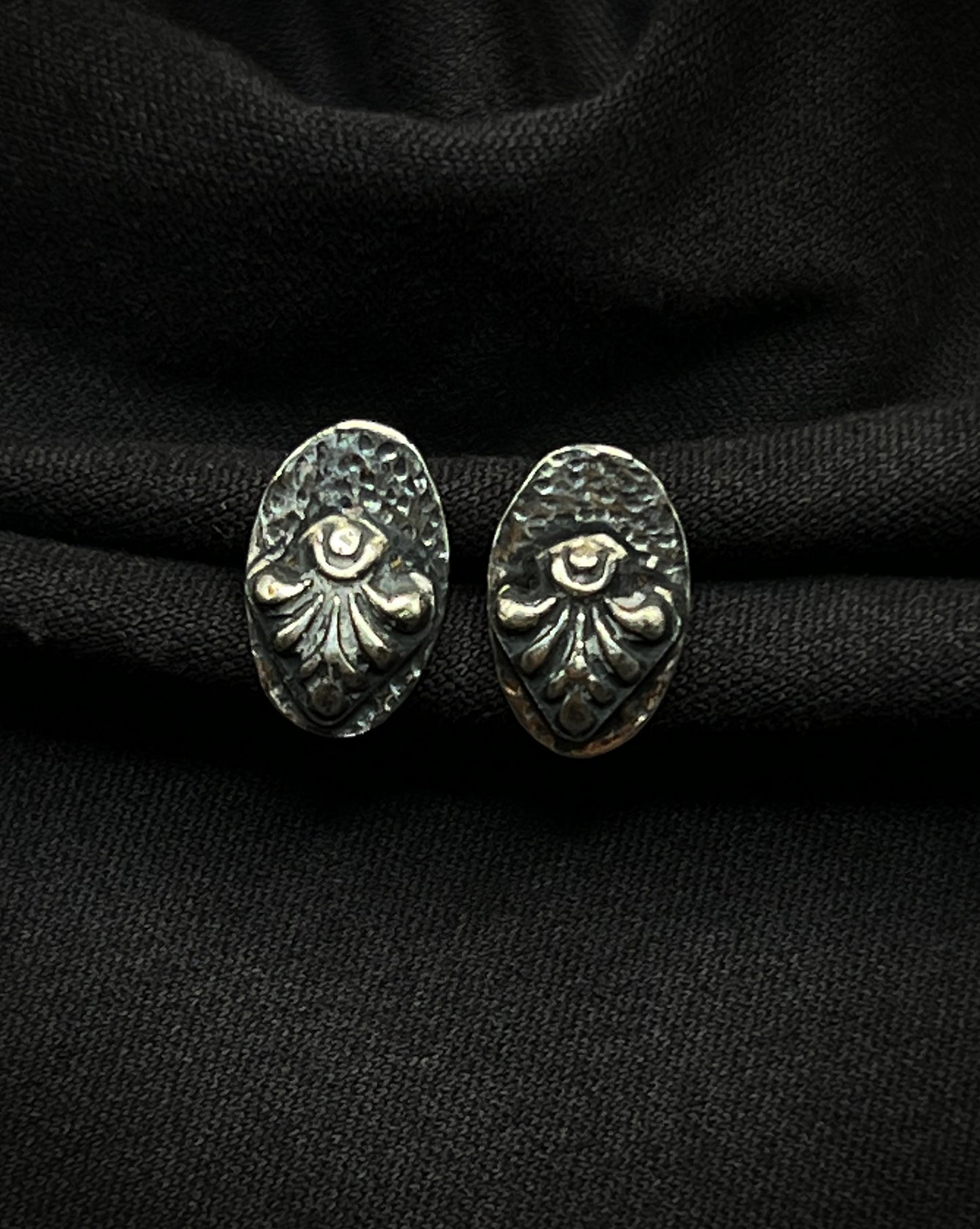A pair of handmade sterling silver stud earrings. Each features a raised, fanned geometric relief centered on a darkened, hammered oval base with a deep charcoal patina.