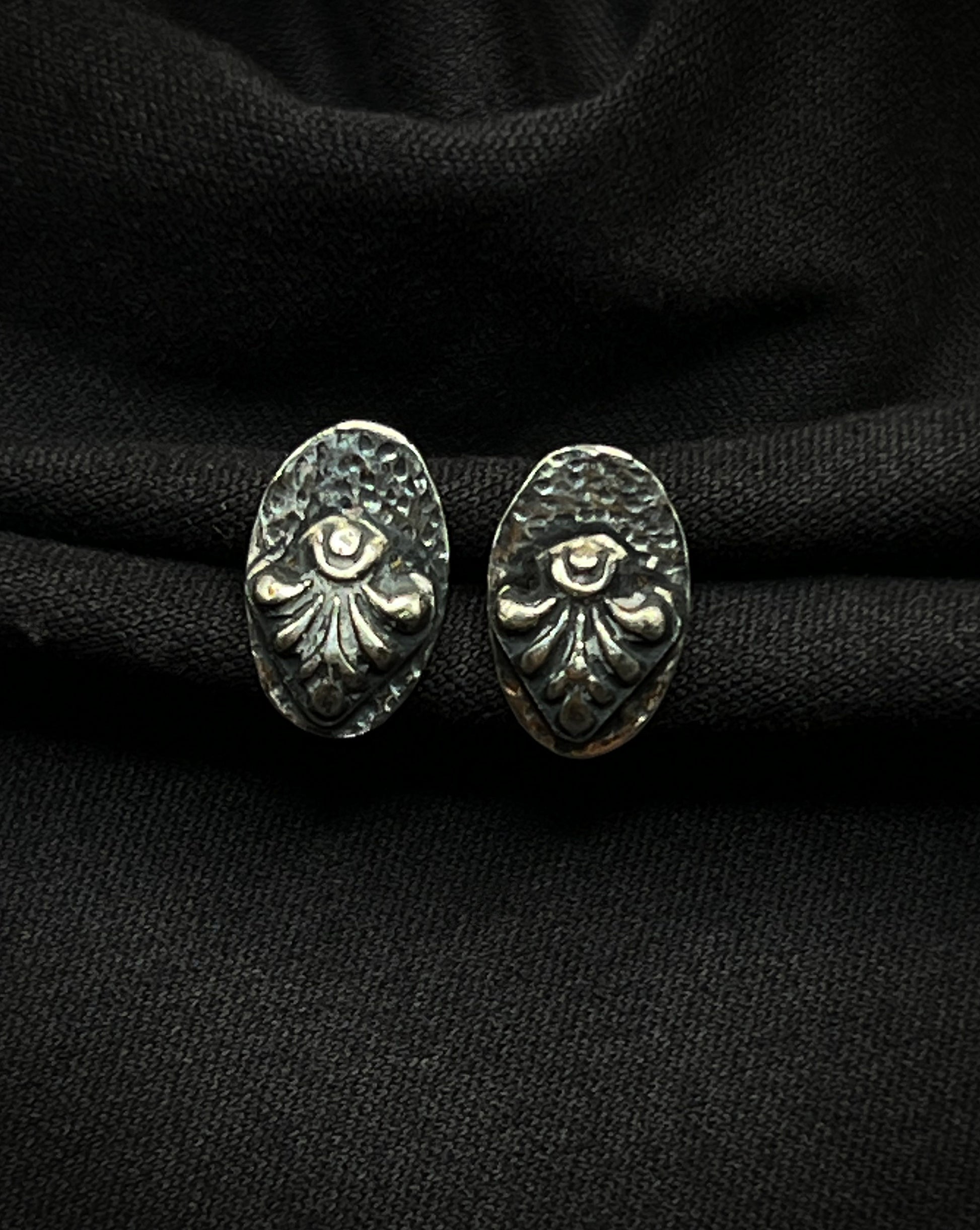 A pair of handmade sterling silver stud earrings. Each features a raised, fanned geometric relief centered on a darkened, hammered oval base with a deep charcoal patina.