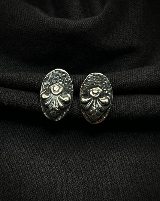 A pair of handmade sterling silver stud earrings. Each features a raised, fanned geometric relief centered on a darkened, hammered oval base with a deep charcoal patina.