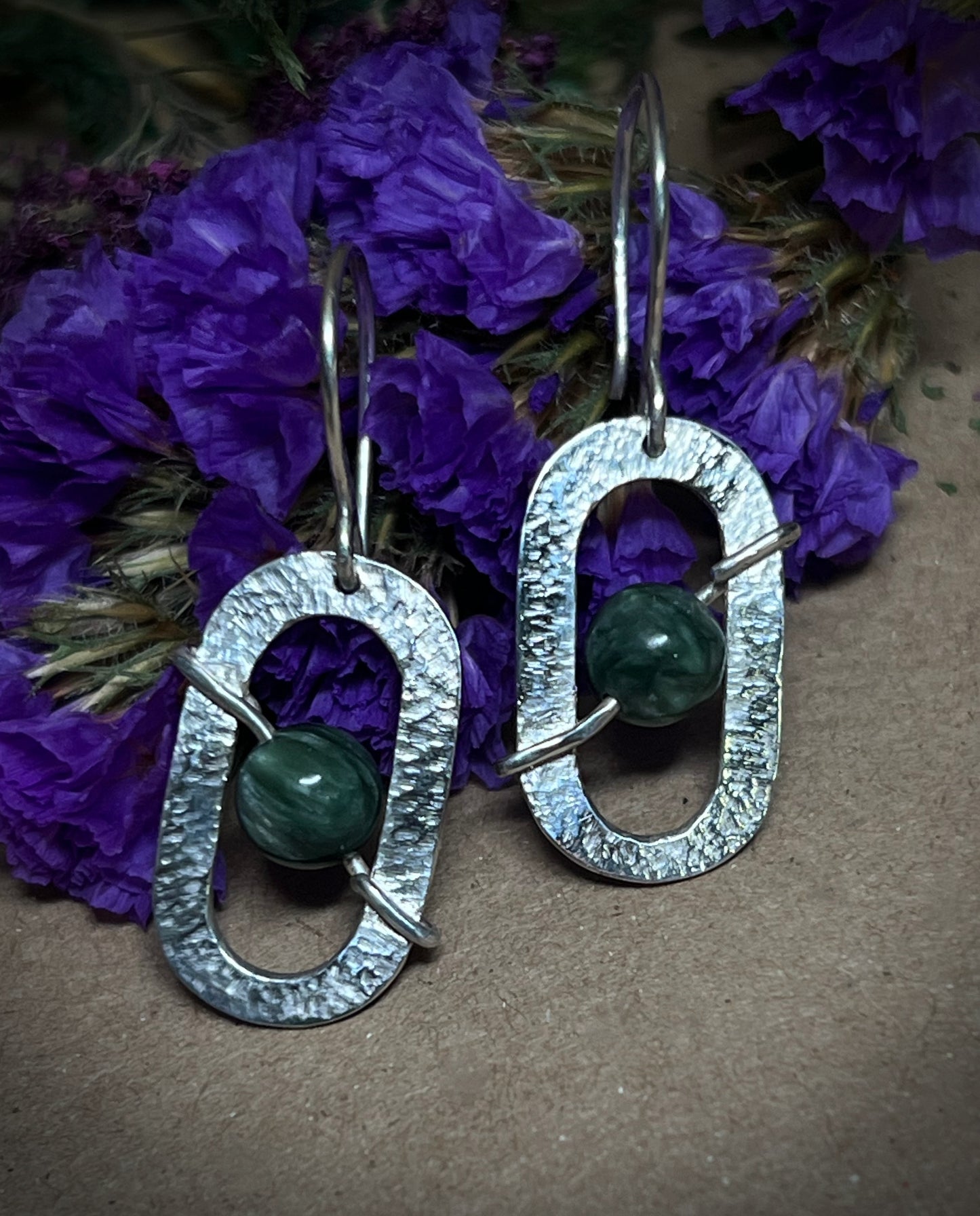 Silver earrings with green beads on a brown surface with purple flowers in the background