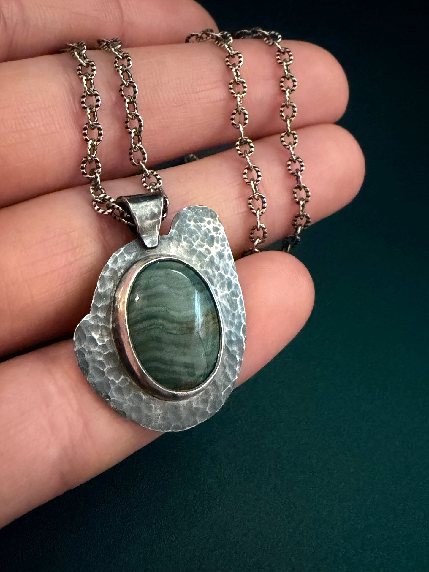Hand holding a silver pendant with a green stone against a green background