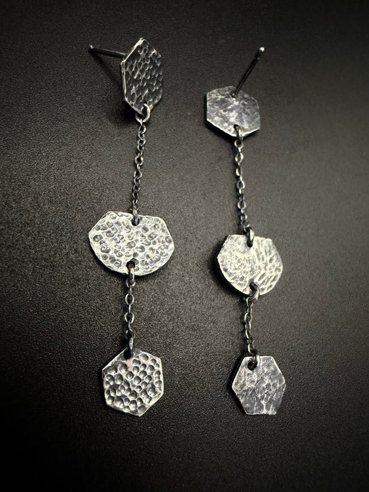 Pair of silver earrings with textured hexagonal pendants on a dark background