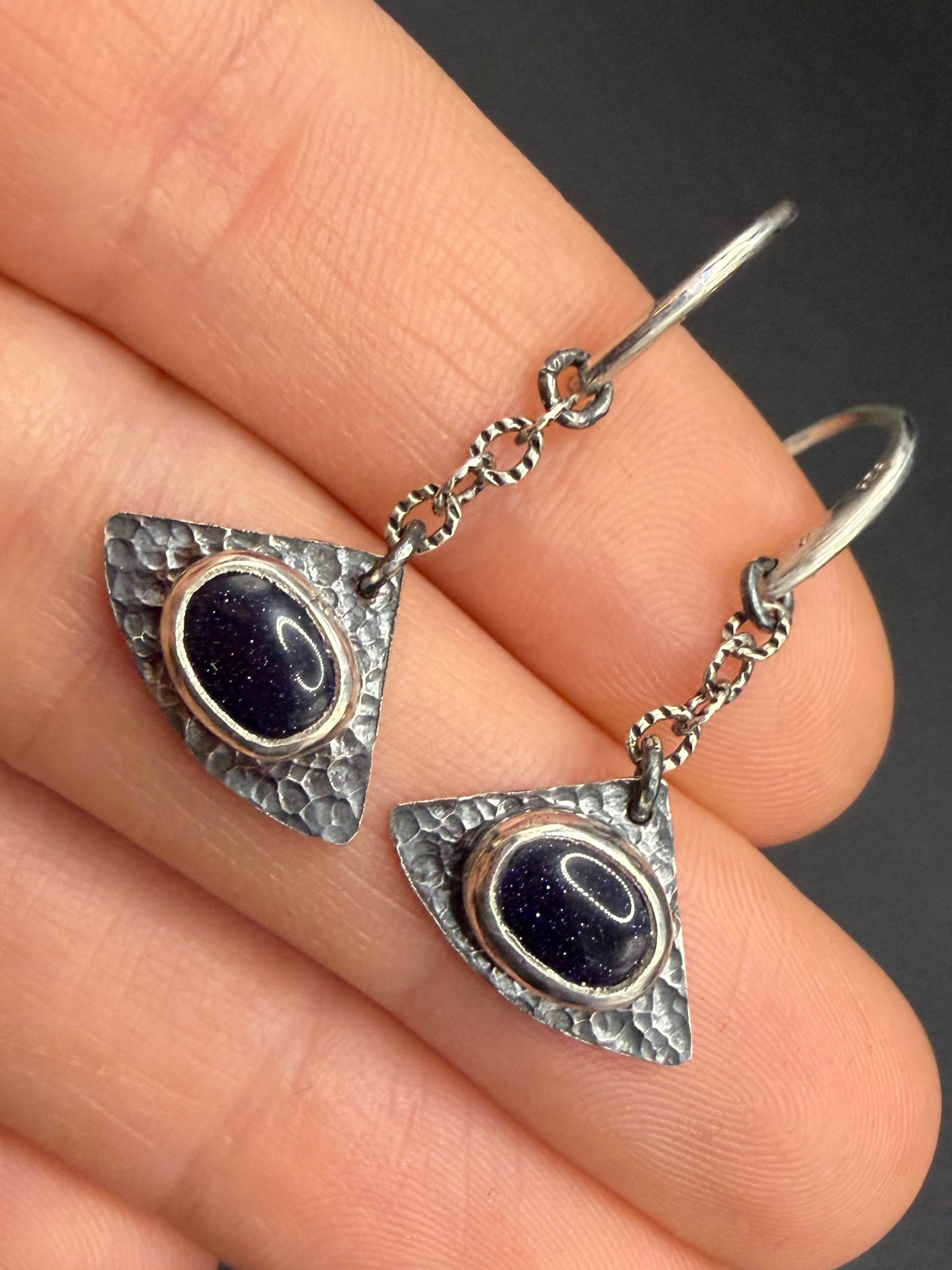 Astraea Drop earrings held in hand to show scale, featuring shimmering blue goldstone galaxy gems set in dark, hammered sterling silver triangular shields.