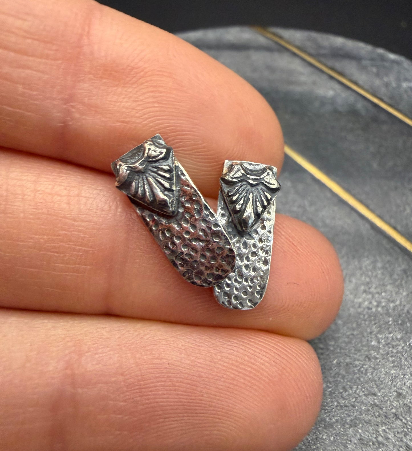 A person holding two Ashborn Root sterling silver studs between their fingers, showcasing the small scale and the contrast between the radiating ridges and the hammered-texture base.