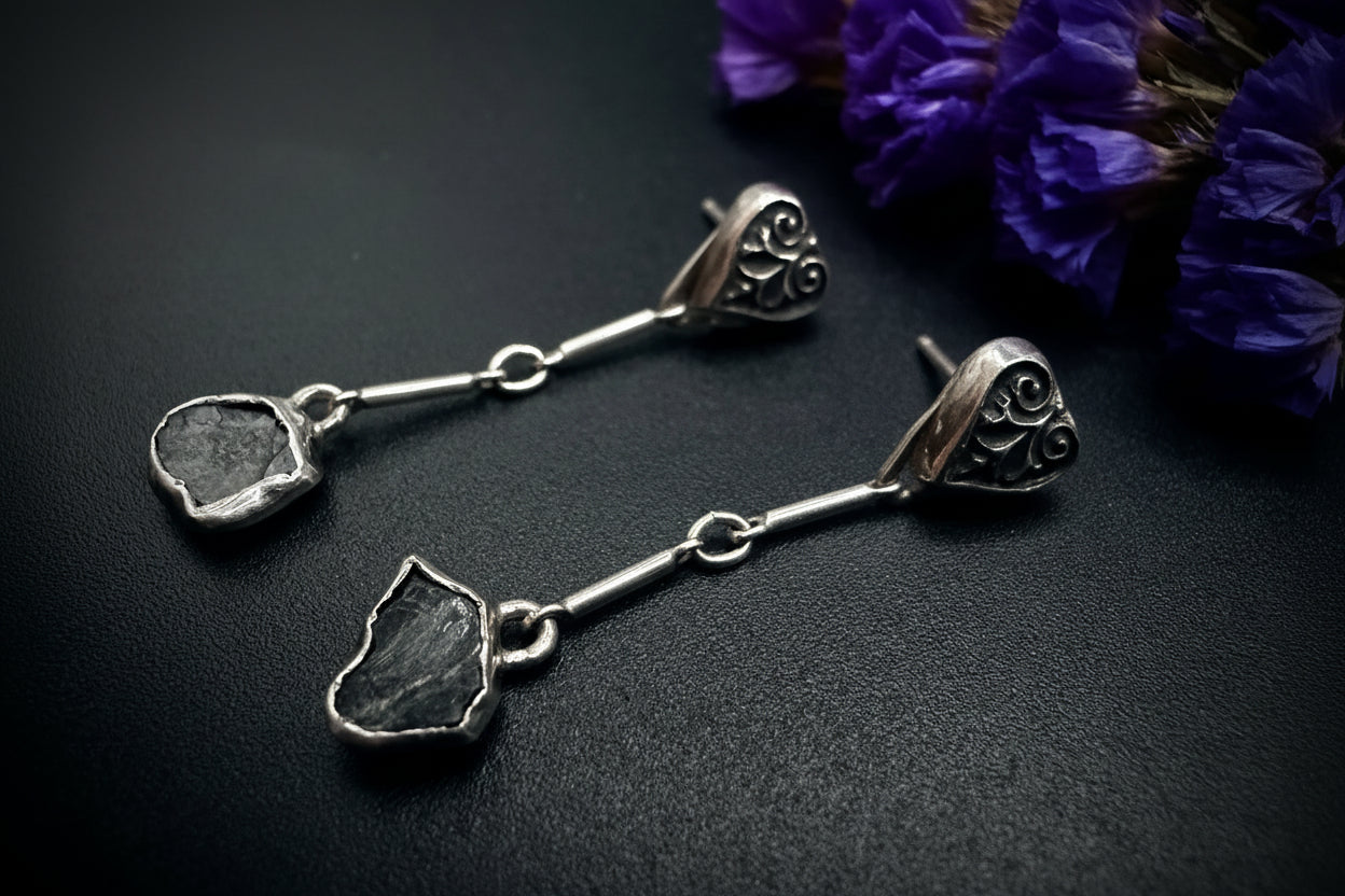 Pair of long sterling silver earrings with sculpted heart-leaf tops and raw, unpolished blue sapphire drops, displayed on a dark textured surface next to purple flowers.