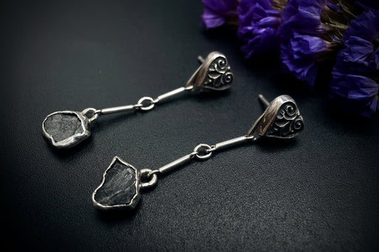 Pair of long sterling silver earrings with sculpted heart-leaf tops and raw, unpolished blue sapphire drops, displayed on a dark textured surface next to purple flowers.