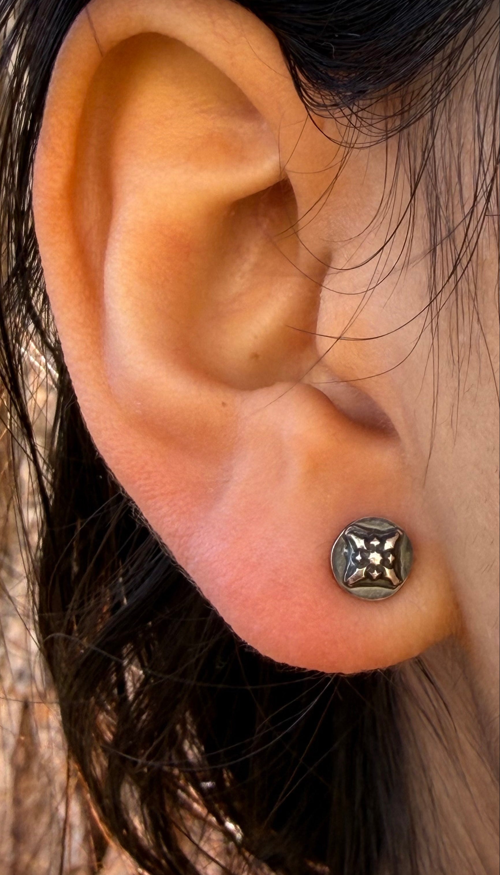 Side view of a model wearing a 7mm Ember Stud from the Ashborn Collection. The sterling silver earring features a polished starburst motif against a dark patina, showcasing its low-profile fit on the earlobe against a natural, sunlit background.