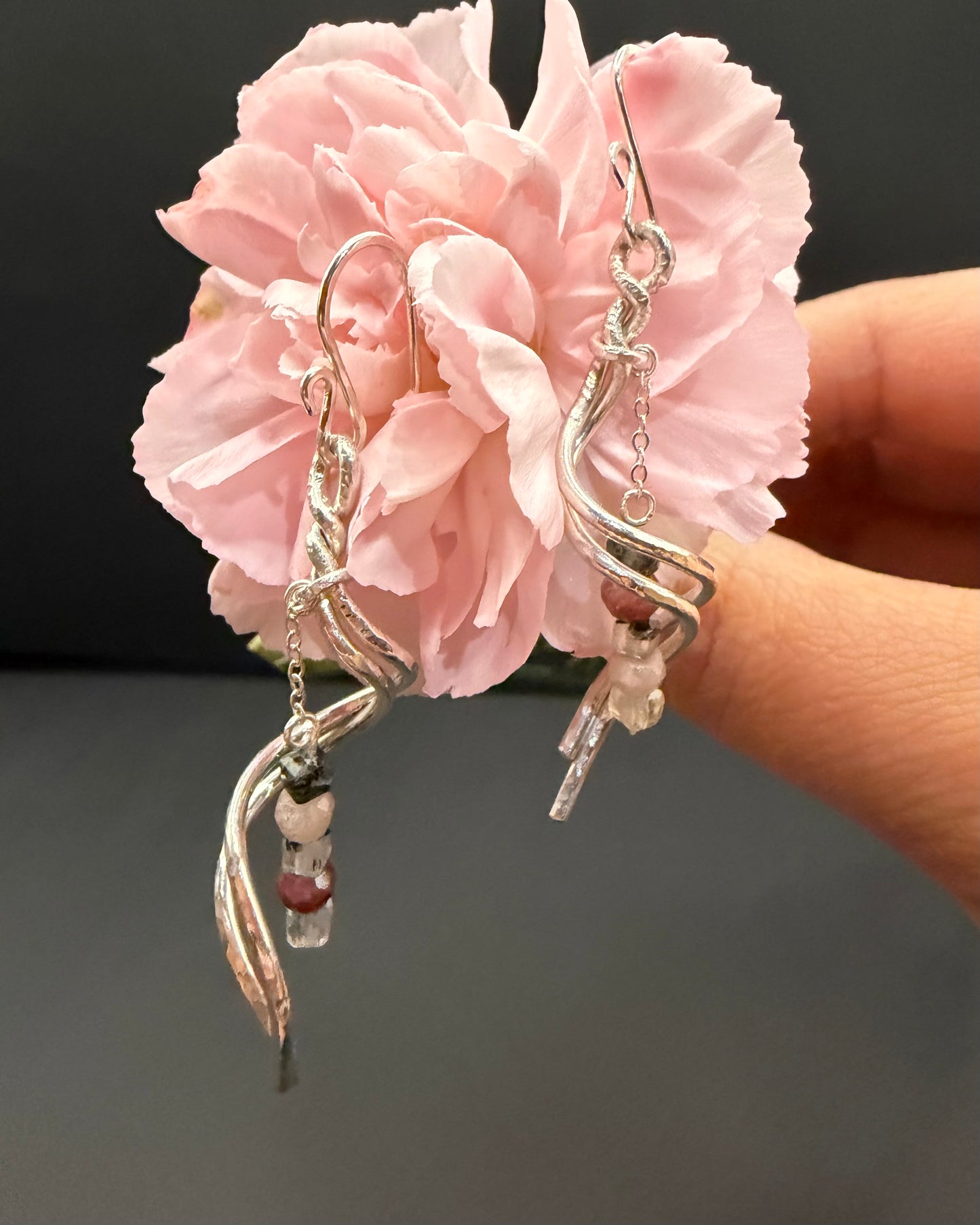 Close-up of the Blushthorn earrings held against a pink carnation, highlighting the ombre bead drop of peach moonstone and green prehnite below the textured silver-tone metal.