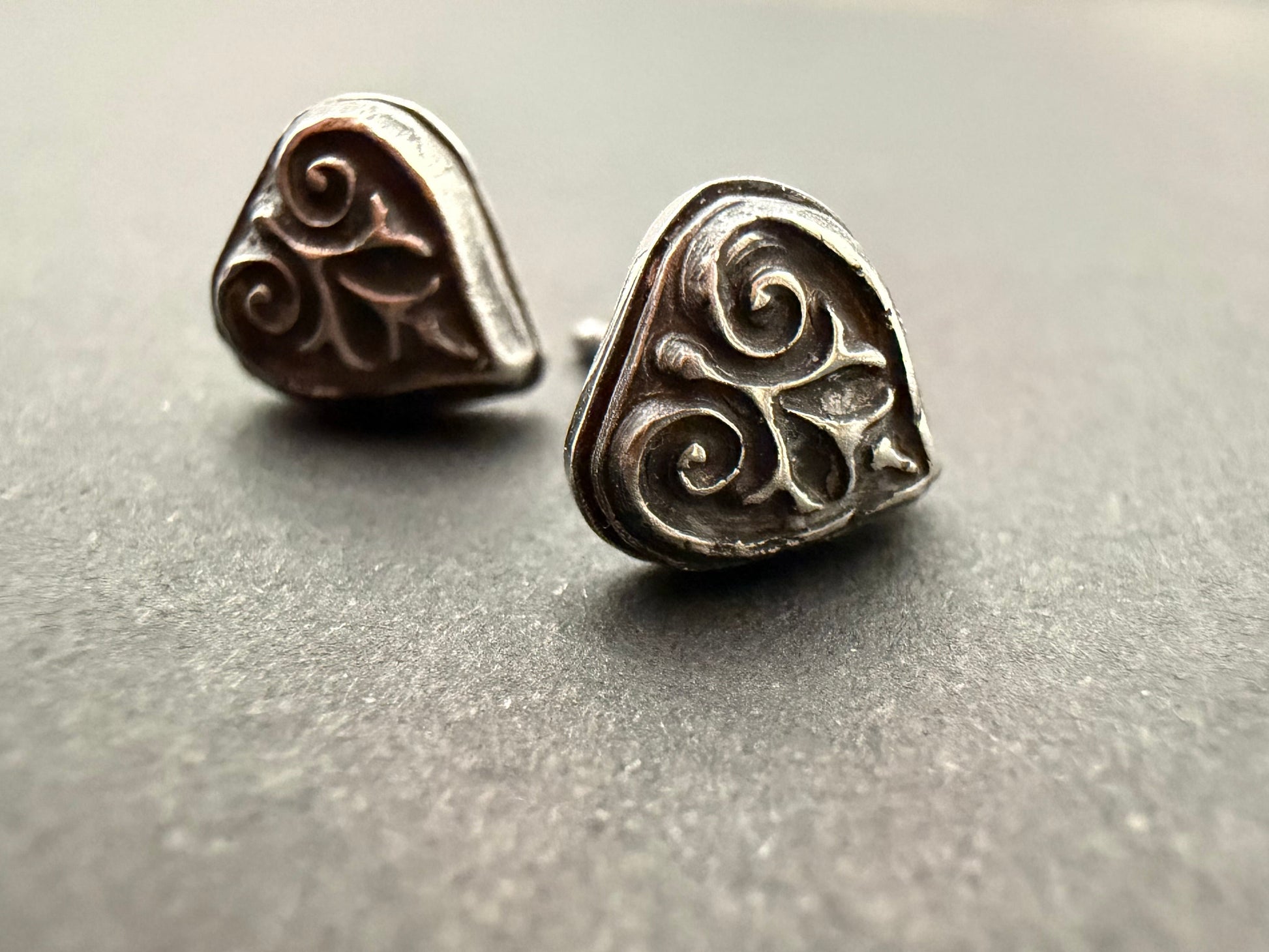 Pair of Ashborn sterling silver studs featuring a raised, triple-scroll design and dark patina, styled for a Dark Academia aesthetic on a textured gray background.