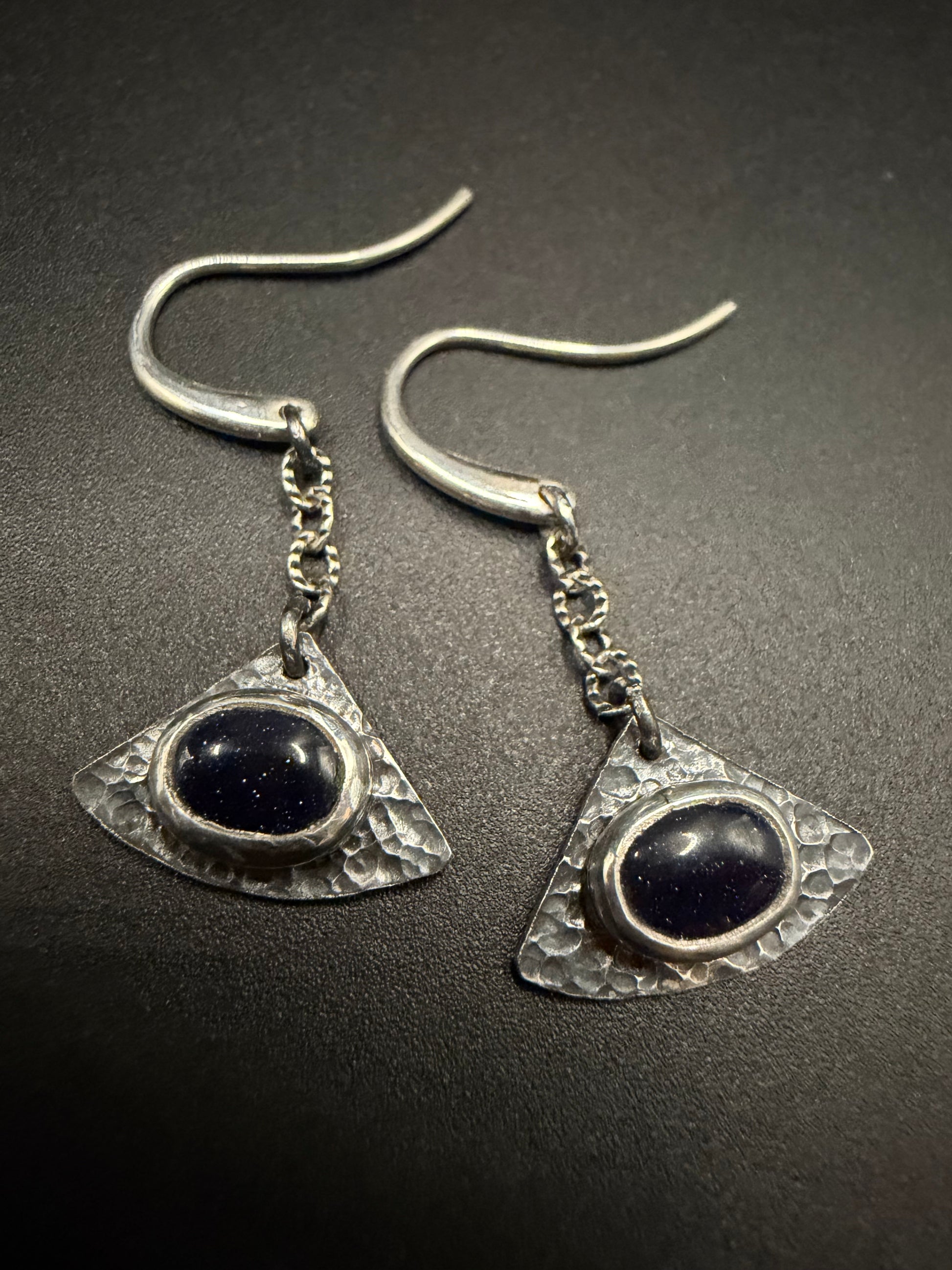 Close-up of Astraea Drop earrings featuring shimmering blue goldstone galaxy stones set in hammered sterling silver triangular shields, styled with a moody Dark Academia vibe.