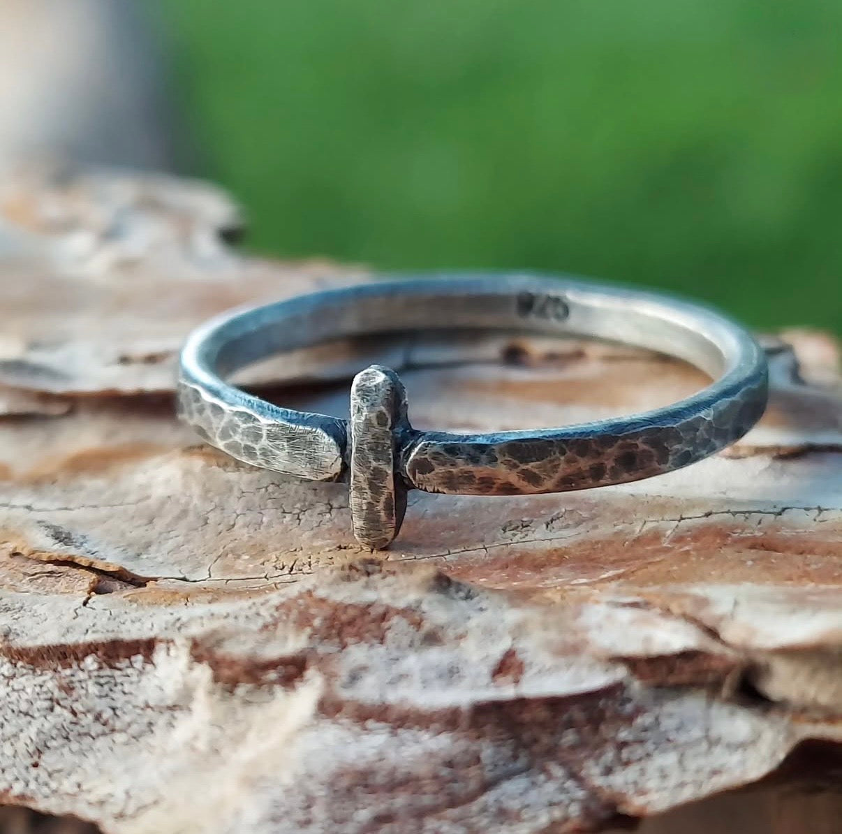 Close-up of a sterling silver minimalist band with a weathered, pitted texture and a single vertical bar design, resting on rough tree bark.
