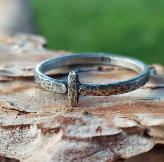 Close-up of a sterling silver minimalist band with a weathered, pitted texture and a single vertical bar design, resting on rough tree bark.