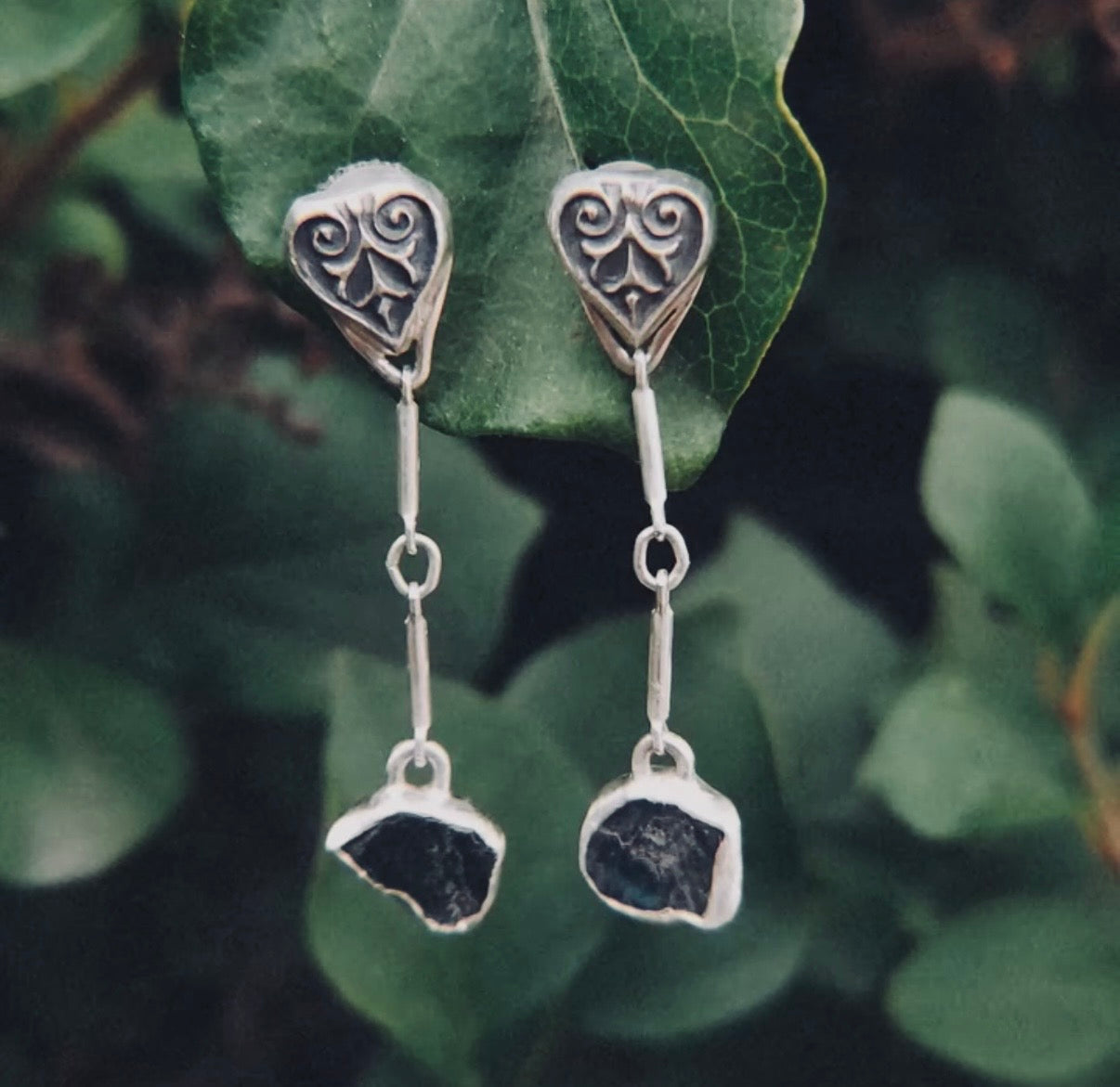 Long sterling silver earrings with sculpted heart-leaf tops and raw, unpolished blue sapphire drops, photographed hanging against dark green leaves.