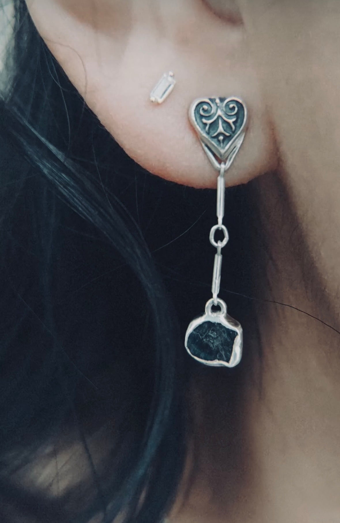A person wearing a long sterling silver drop earring featuring a sculpted heart-leaf top and a raw, unpolished blue sapphire stone suspended by a delicate silver link.