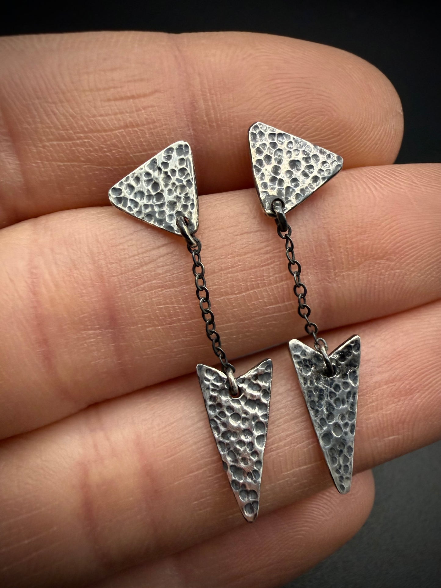 Hammered sterling silver arrow earrings with an antiqued patina, held between fingers to show scale against a dark background.