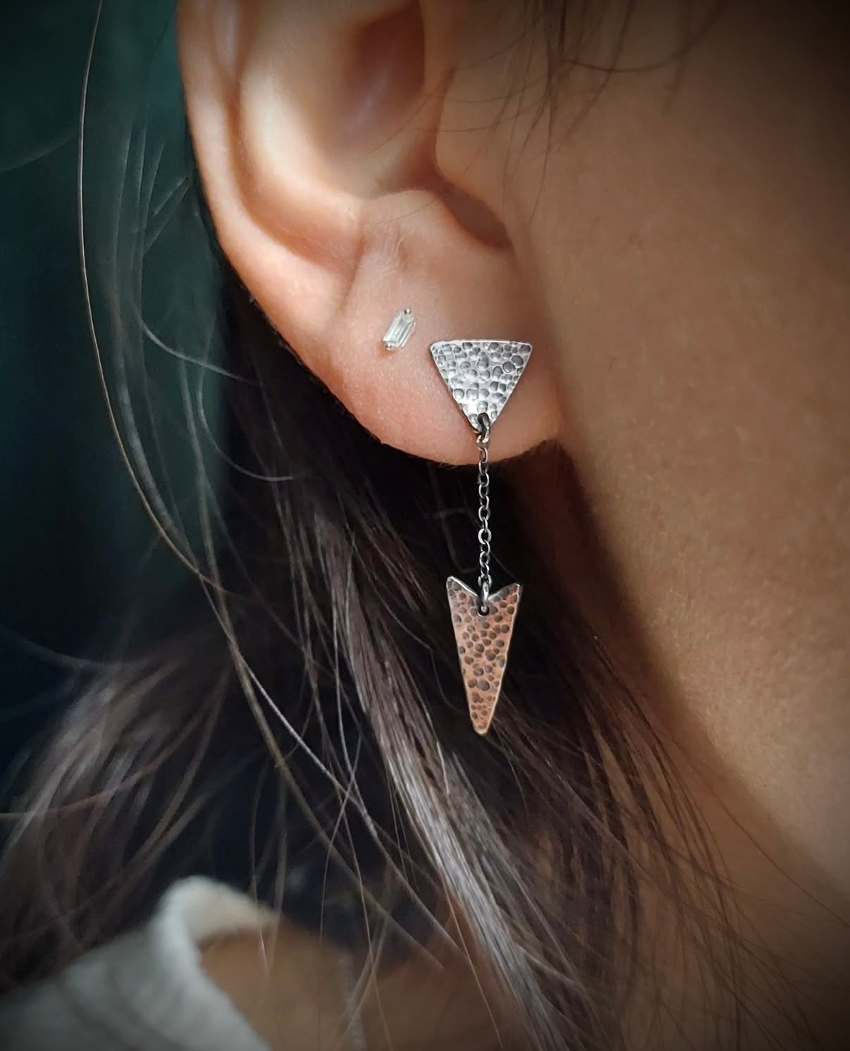 On-ear view of a hammered sterling silver arrow earring. The textured triangle stud connects to a dangling arrowhead by a delicate silver chain, showing the drop length against the jawline.