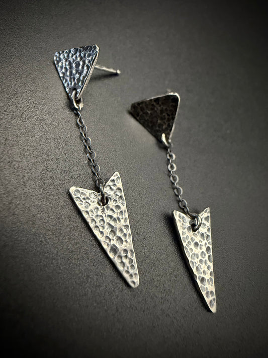 Long hammered sterling silver arrow earrings with a dark antiqued patina and dangling chain detail, shown against a moody dark background.