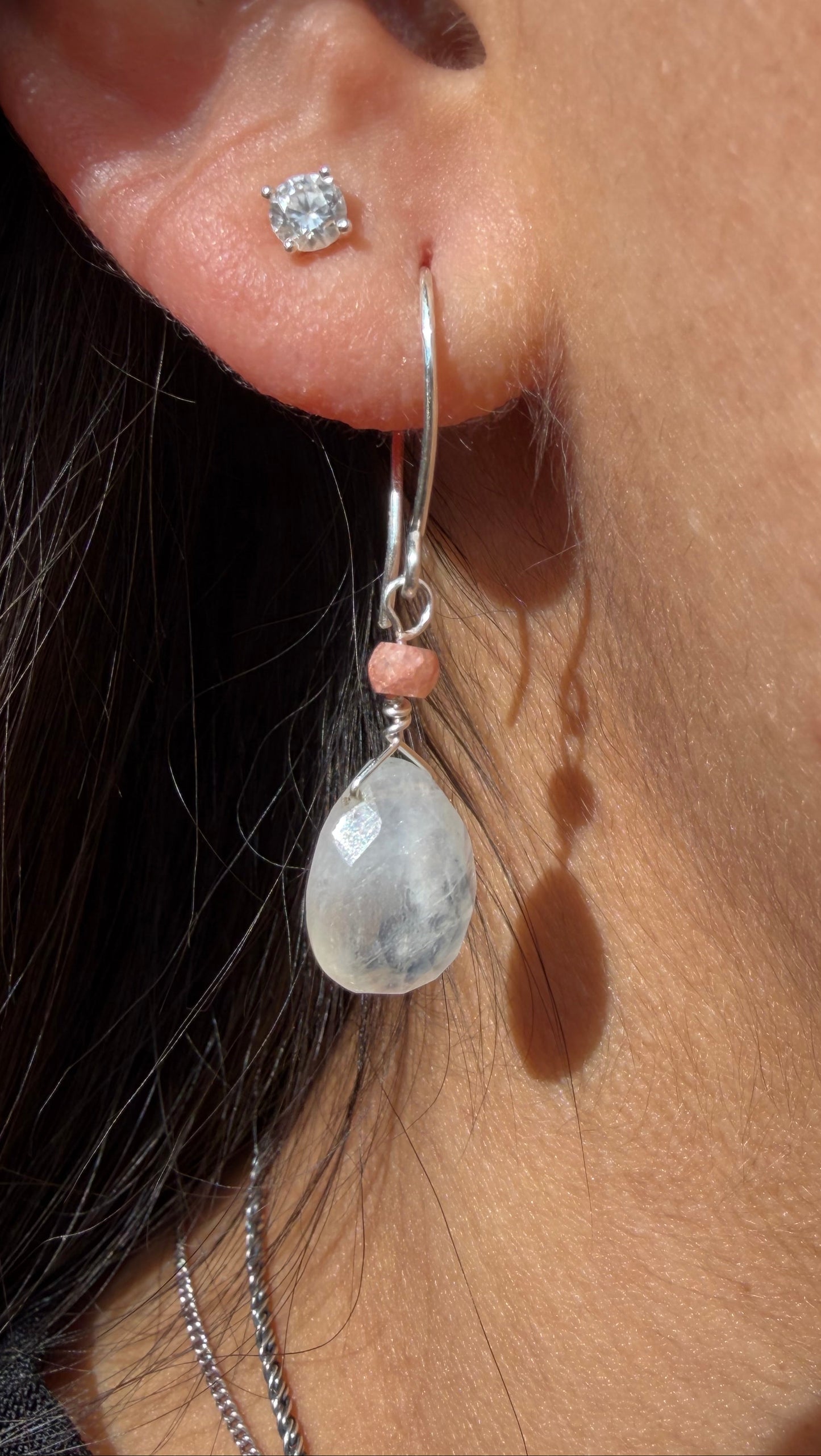 Close-up of a person wearing Lotus Drops earrings featuring a faceted white moonstone teardrop and a small peach moonstone bead on a sterling silver wire.