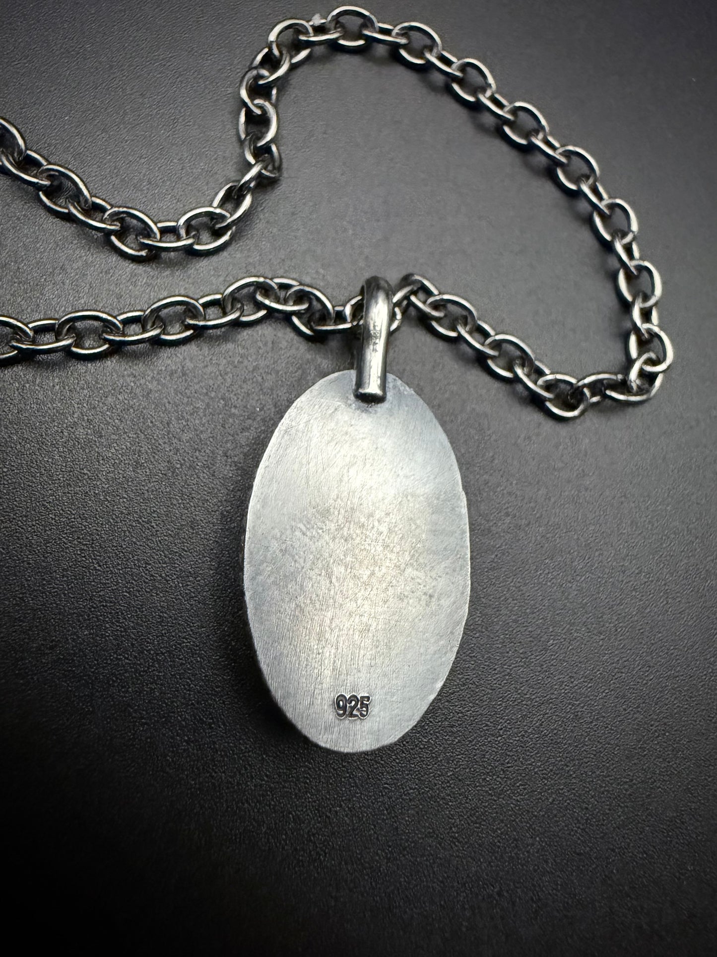 The back of a sterling silver oval pendant showing a hand-brushed matte finish and a "925" hallmark stamp, attached to a sturdy oxidized silver chain