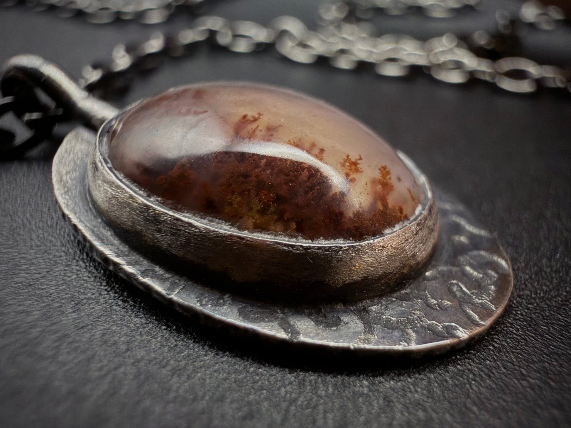 Close-up of an oval red Plume Agate stone with natural moss-like inclusions, secured in a hand-textured sterling silver bezel with a dark antiqued patina.