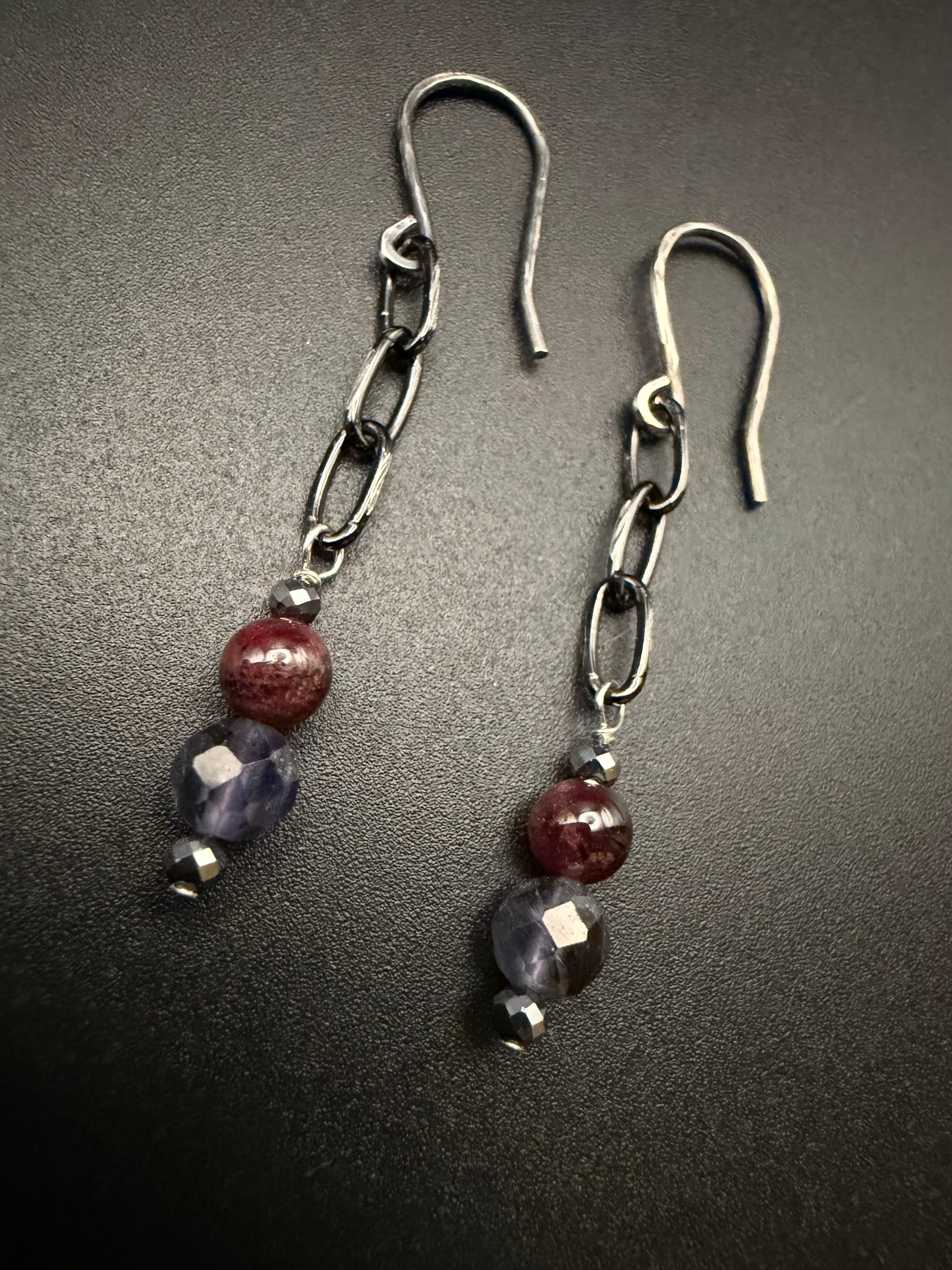 Handcrafted sterling silver earrings featuring a chain drop with a vertical stack of metallic hematite, deep red garnet, and faceted purple iolite beads on a dark background.