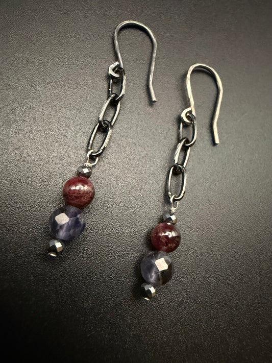 Handcrafted sterling silver earrings featuring a chain drop with a vertical stack of metallic hematite, deep red garnet, and faceted purple iolite beads on a dark background.