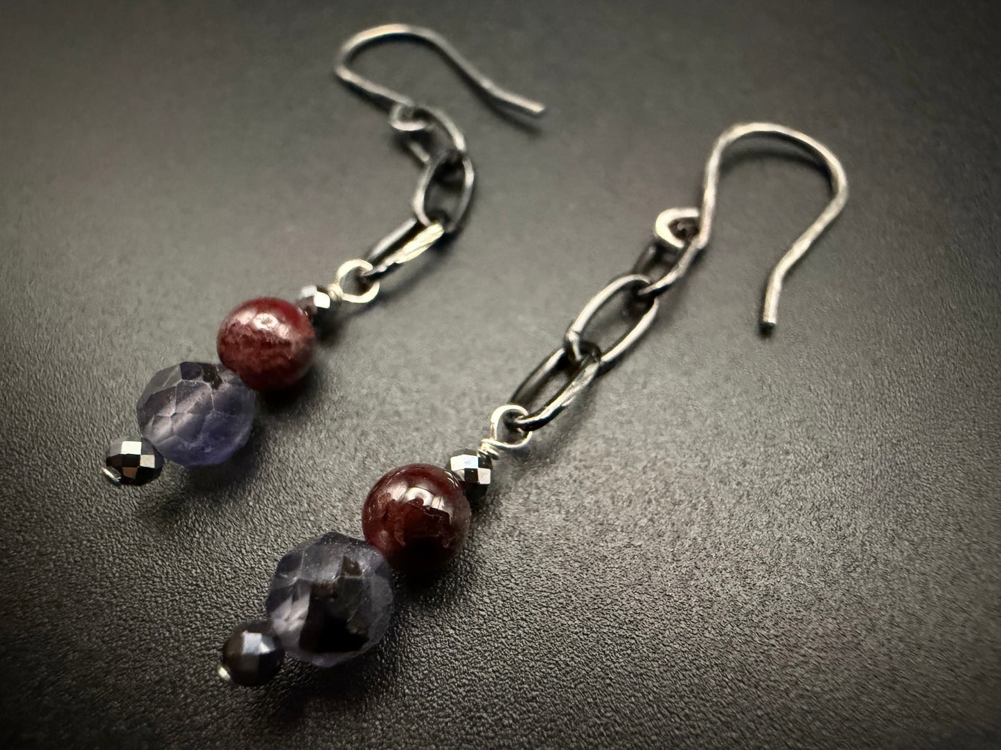 Handcrafted sterling silver earrings featuring a vertical stack of metallic hematite, deep red garnet, and faceted purple iolite beads on a dark, textured background.