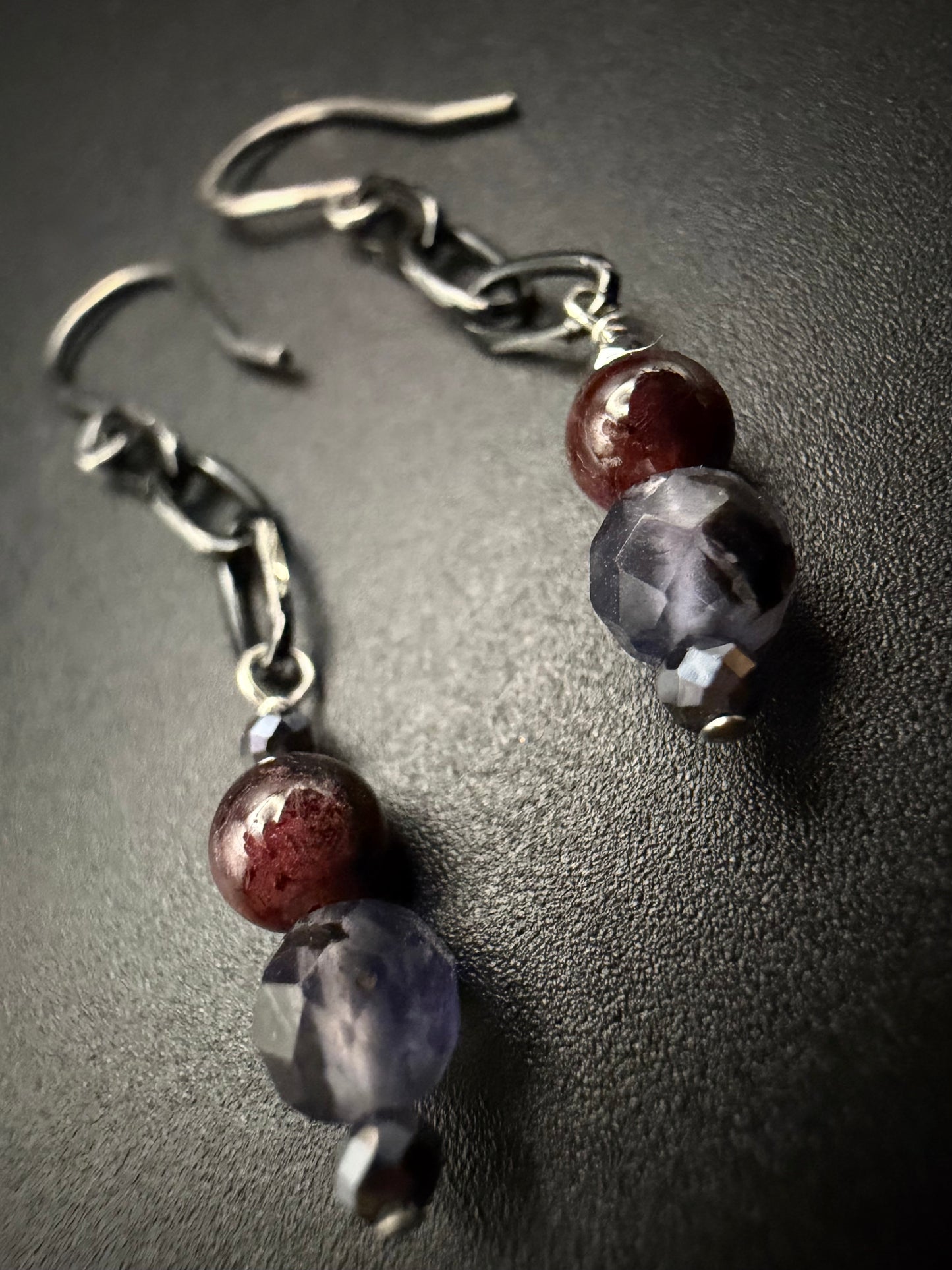 A pair of handcrafted sterling silver drop earrings featuring metallic hematite, deep red garnet, and faceted purple iolite beads on a dark textured surface.