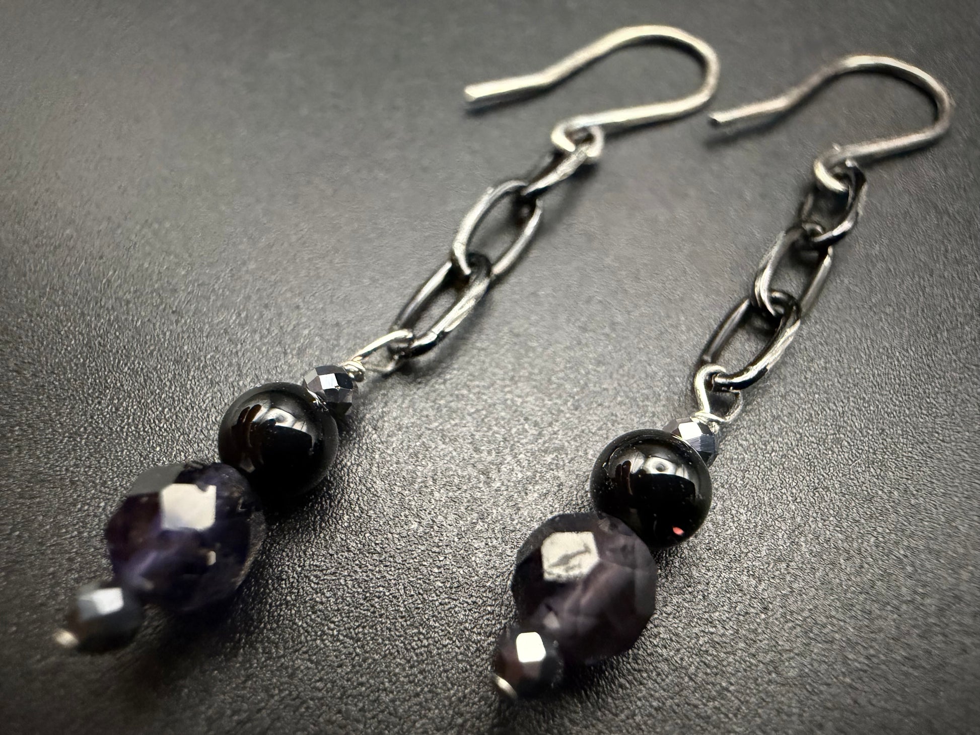 A pair of handcrafted sterling silver drop earrings featuring faceted purple iolite, polished black onyx, and metallic hematite beads on a silver chain.