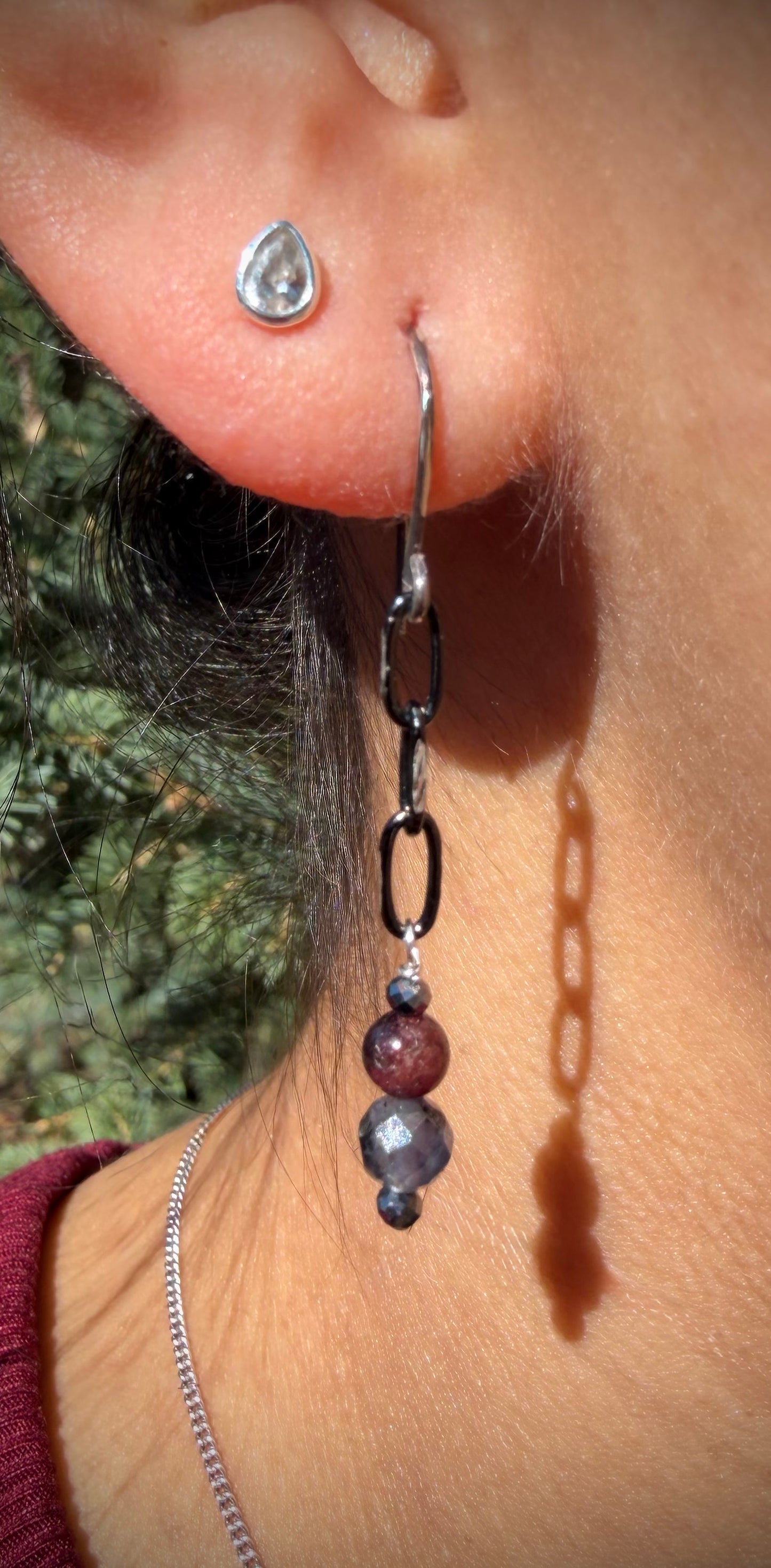 Close-up of a person wearing the Amara Drops: handcrafted sterling silver earrings featuring a dark chain with a vertical stack of hematite, garnet, and iolite beads.