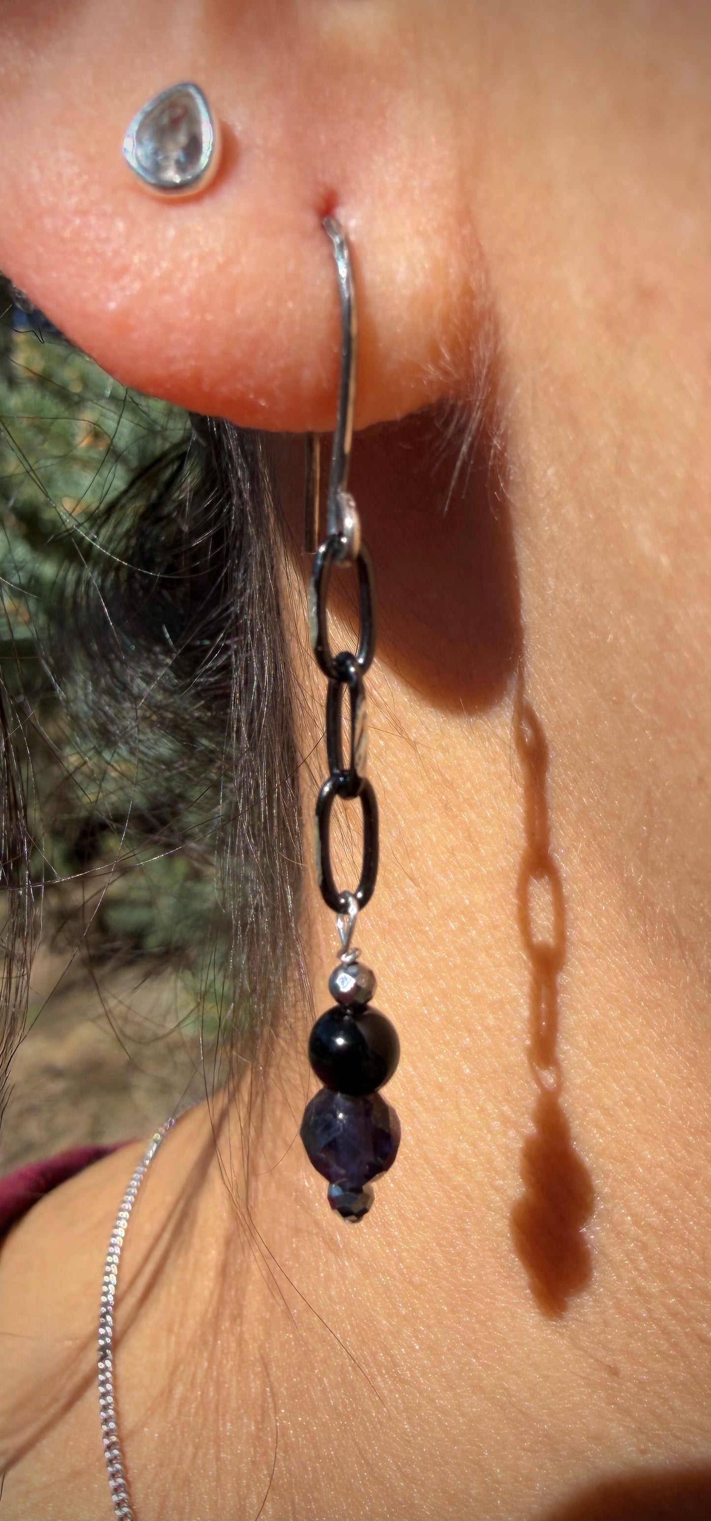 Close-up of a person wearing a handcrafted sterling silver earring featuring a dark patina chain and a vertical stack of onyx, iolite, and hematite beads.