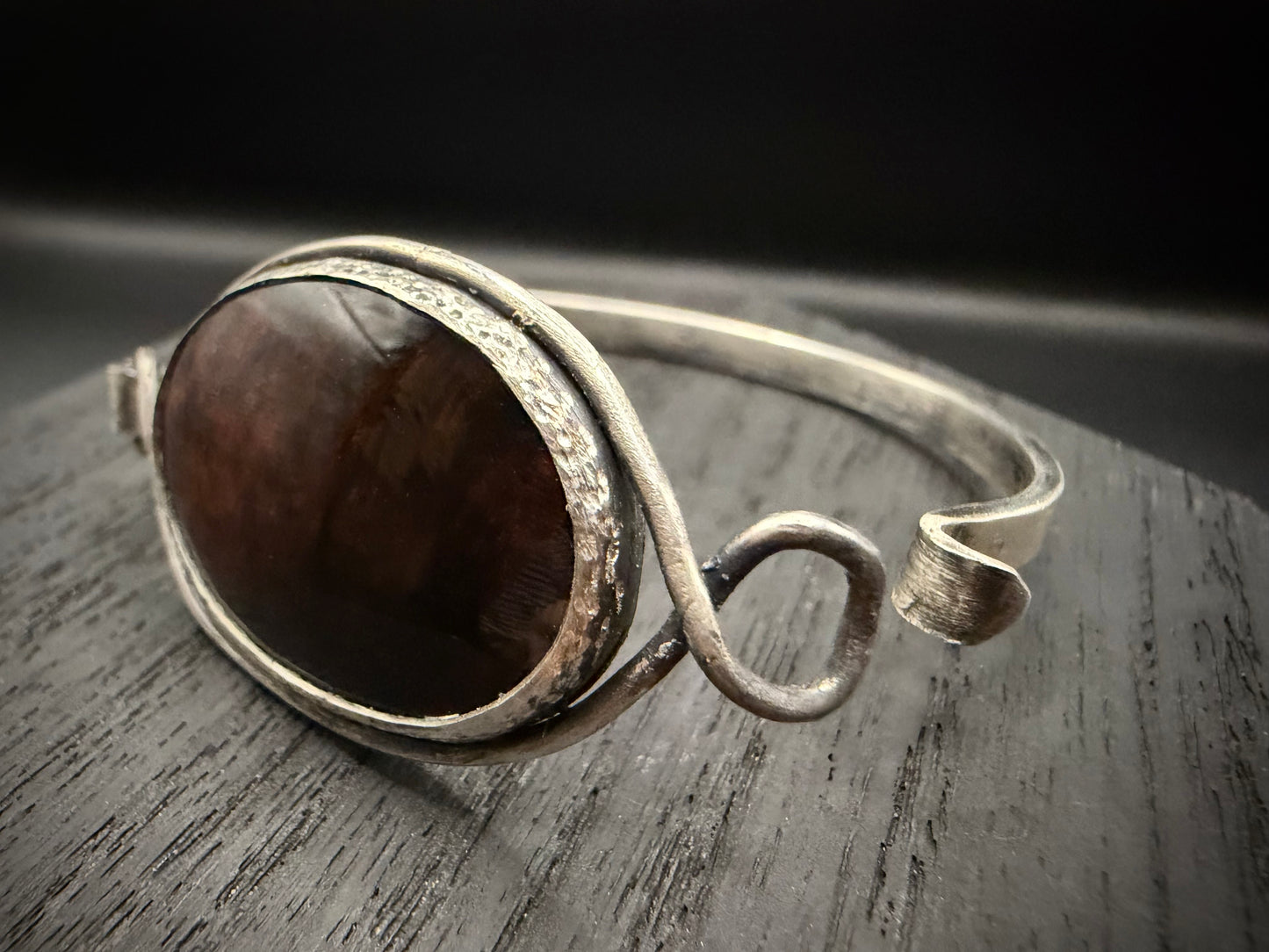 A side view of a grungy, forest-inspired silver cuff showing the intentional rough-hewn texture and the unique reddish-brown inclusions of the mahogany obsidian.