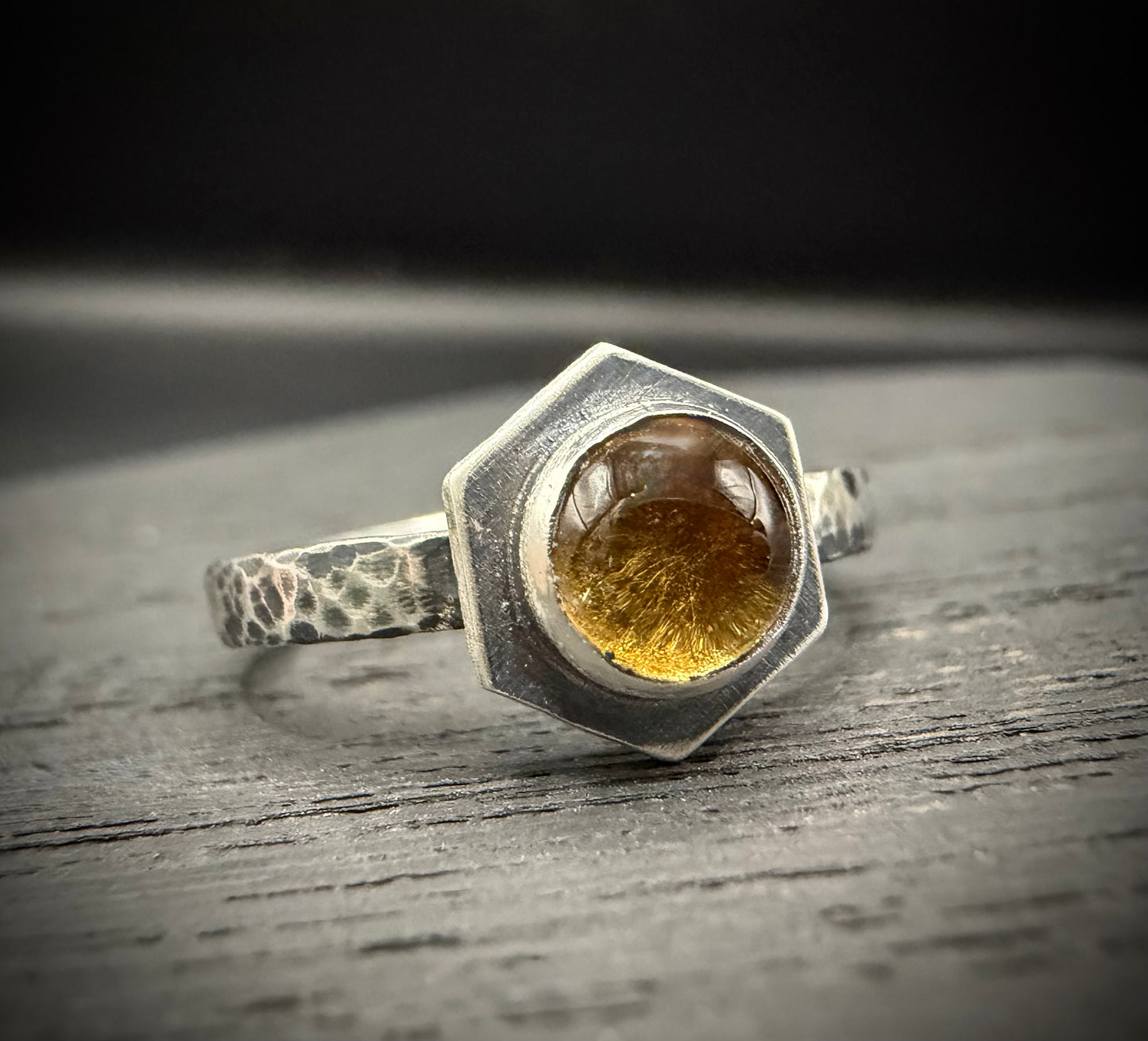 Solaire Ring featuring a hexagonal citrine stone set in a heavy, hand-hammered silver band, displayed on a rustic, dark wooden surface.