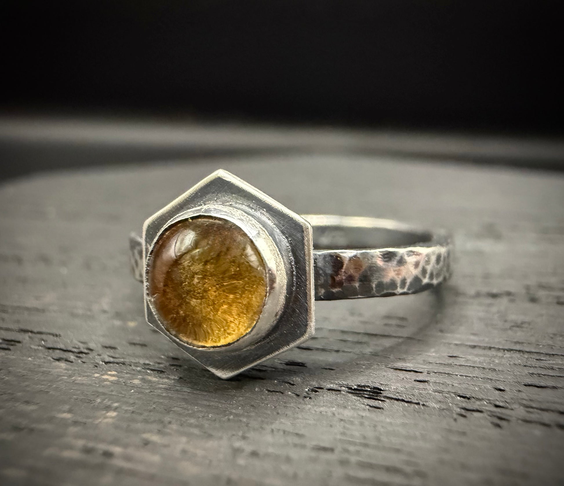 Close-up of the Solaire Ring featuring a hexagonal citrine stone set in a heavy, hand-hammered sterling silver band with an oxidized, moody finish on a dark rustic surface.