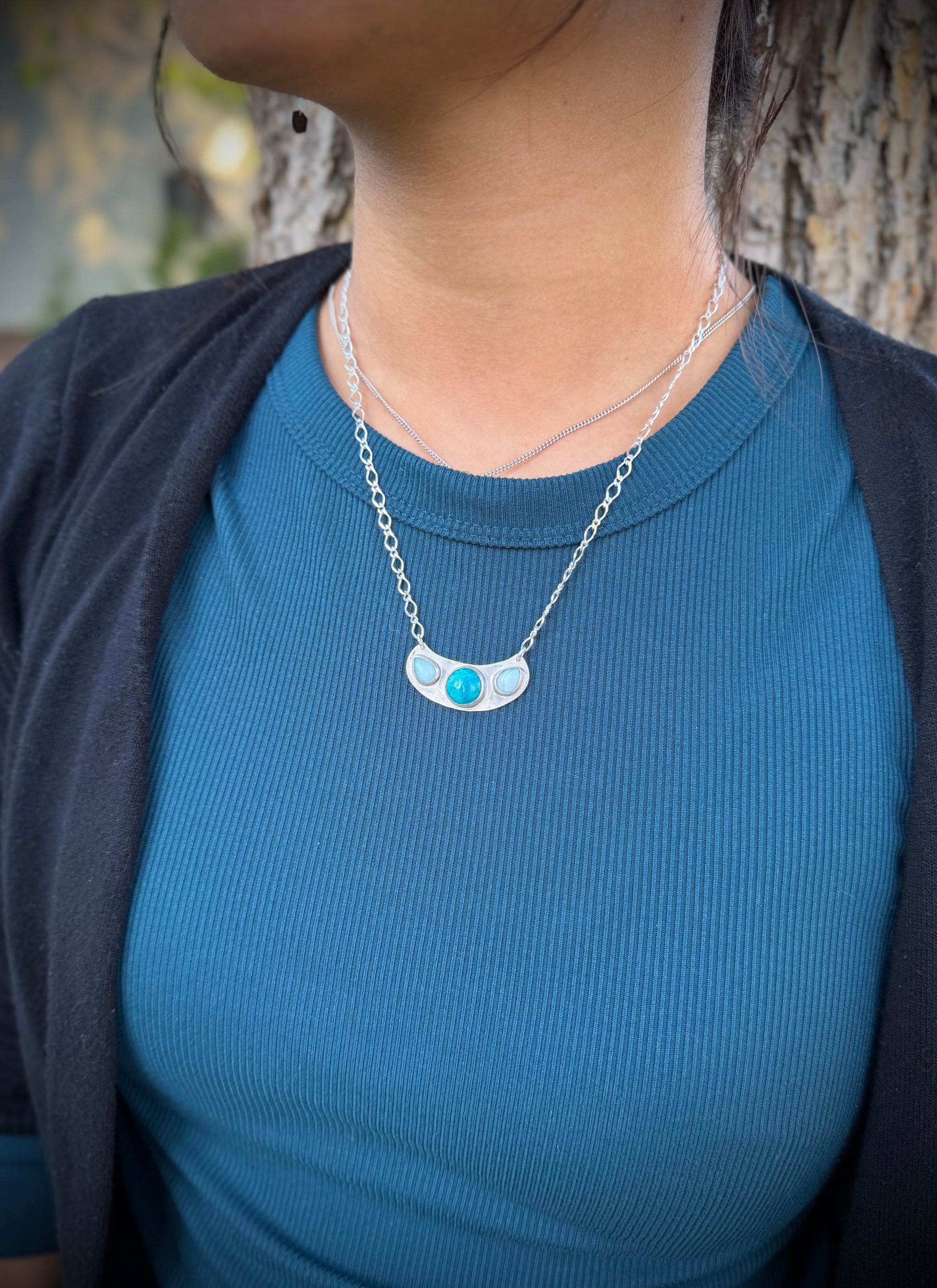 A person wearing the handcrafted sterling silver Marisol Pendant over a blue ribbed shirt and a dark cardigan. The image highlights how the tiered pendant—featuring its teal Chrysocolla and light blue Larimar stones—sits against the chest, showcasing the scale of the piece and the way the long-link silver chain layers naturally.